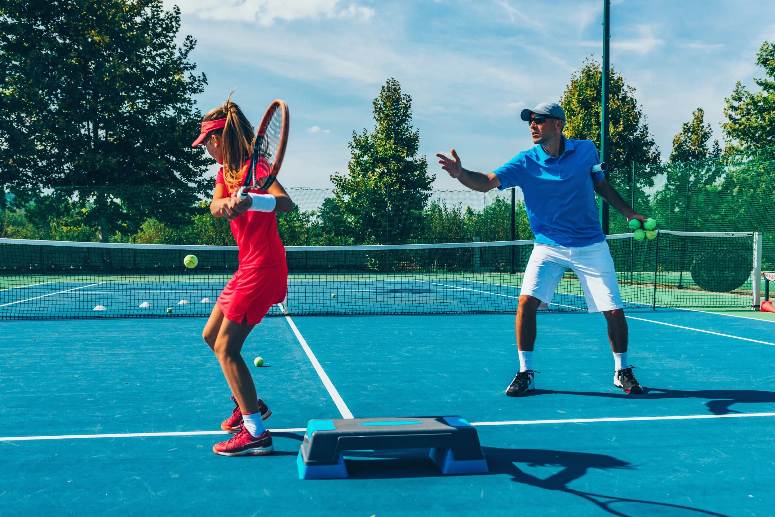 A man coaching a young girl on a tennis court, with the girl preparing to hit a tennis ball while the coach gestures with his hand, under a blue sky and surrounded by trees.