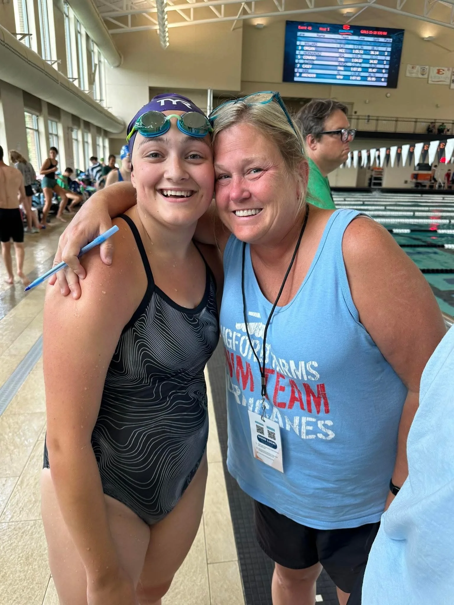 A young female swimmer in a black swimsuit and swimming goggles hugs a smiling older woman wearing a blue tank top and sunglasses at an indoor swimming pool.