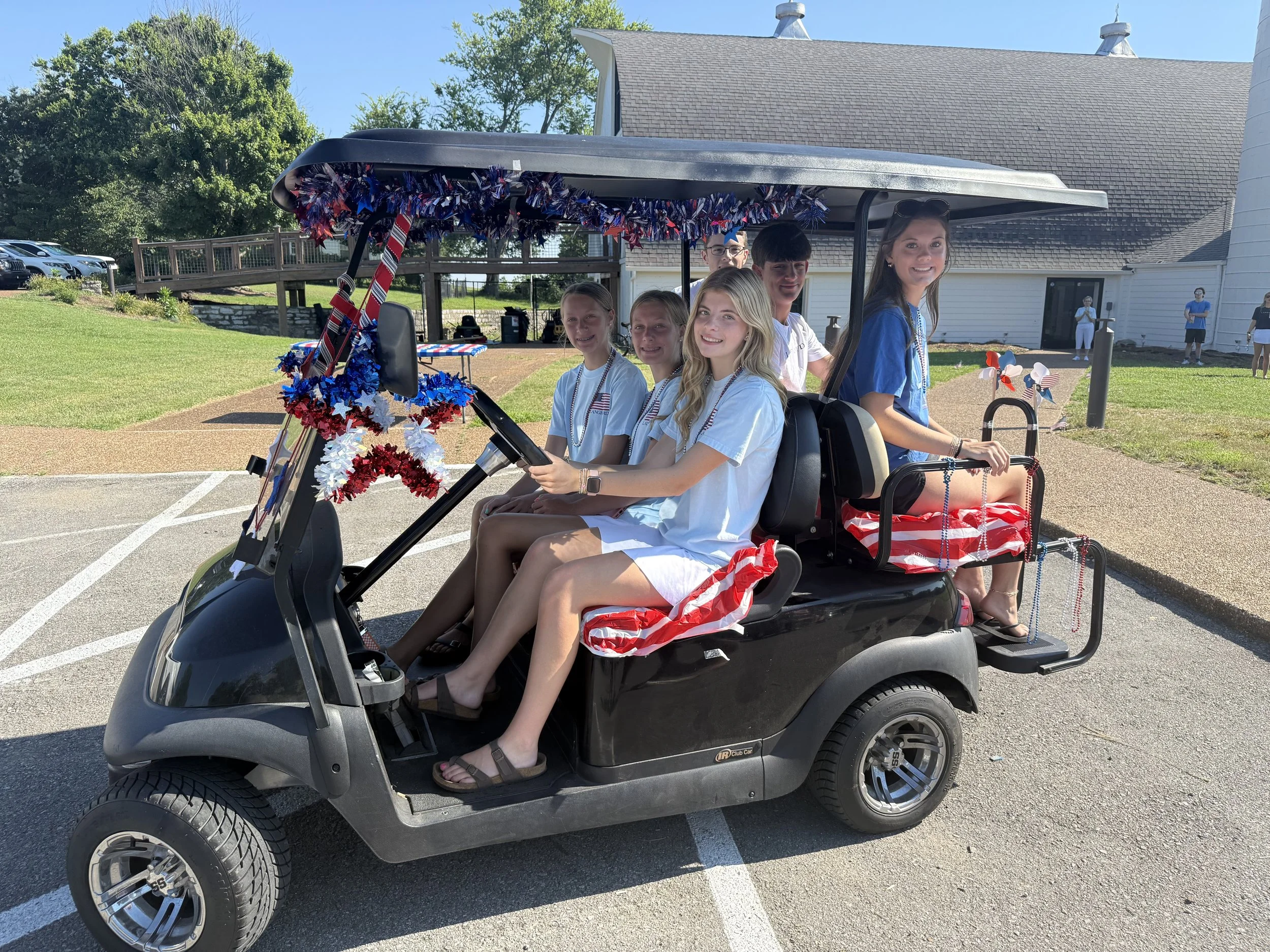 A group of young people sitting in a decorated golf cart on a sunny day, with a building and trees in the background, celebrating a patriotic event.
