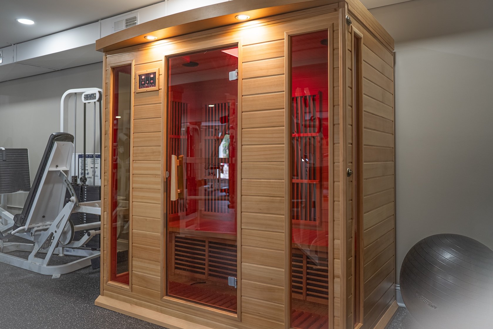 A wooden infrared sauna with glass doors and red interior lighting in a gym or wellness center. Exercise equipment and a stability ball are visible nearby.