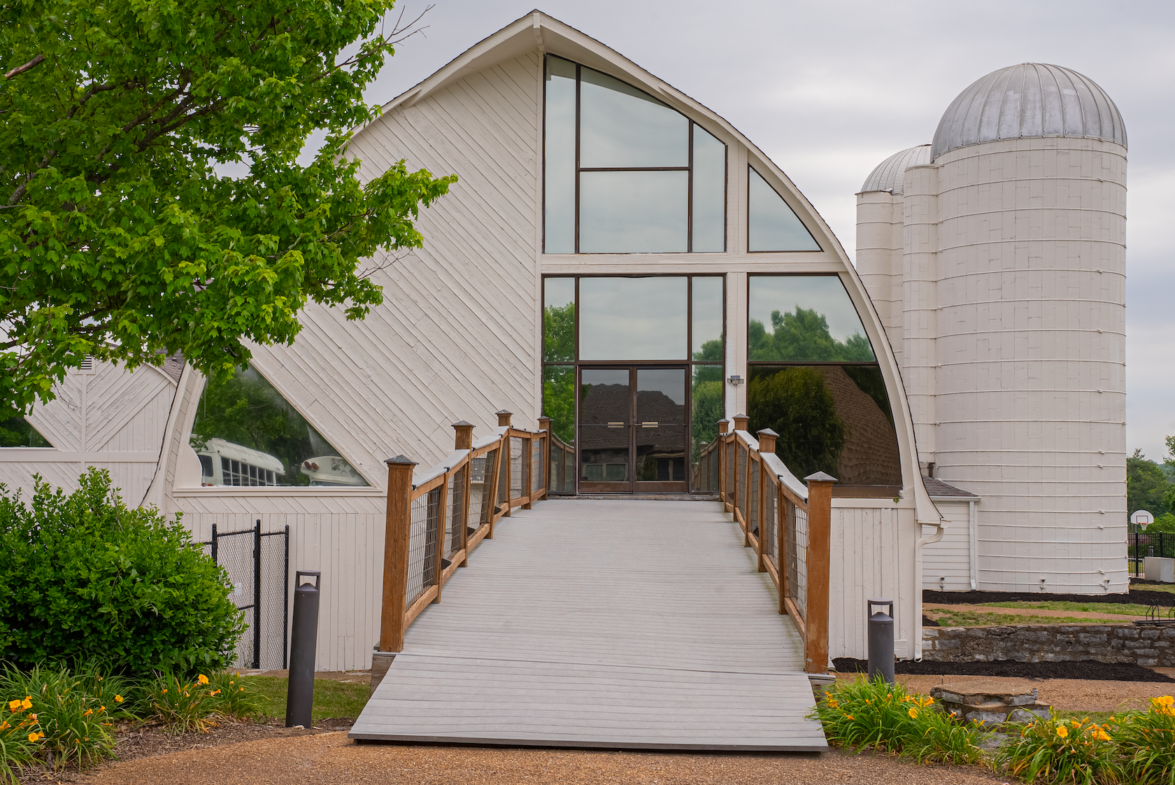A modern building with a boat-shaped roof and large glass windows, surrounded by green trees and plants, with a wooden ramp leading to the entrance.