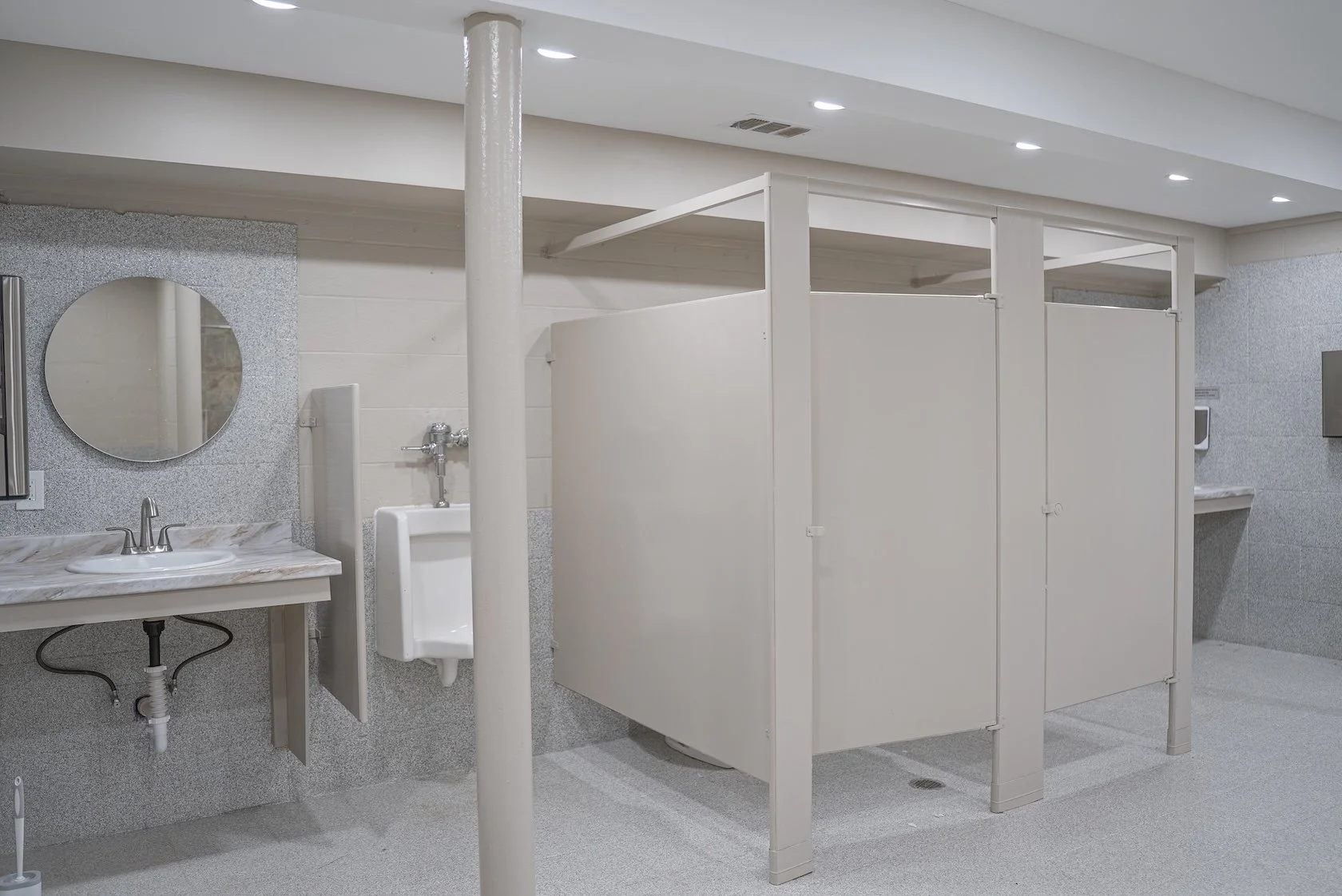 Bathroom with multiple stalls, a sink with a marble countertop, a mirror, and a urinal. The walls are tiled, and the ceiling has recessed lighting.