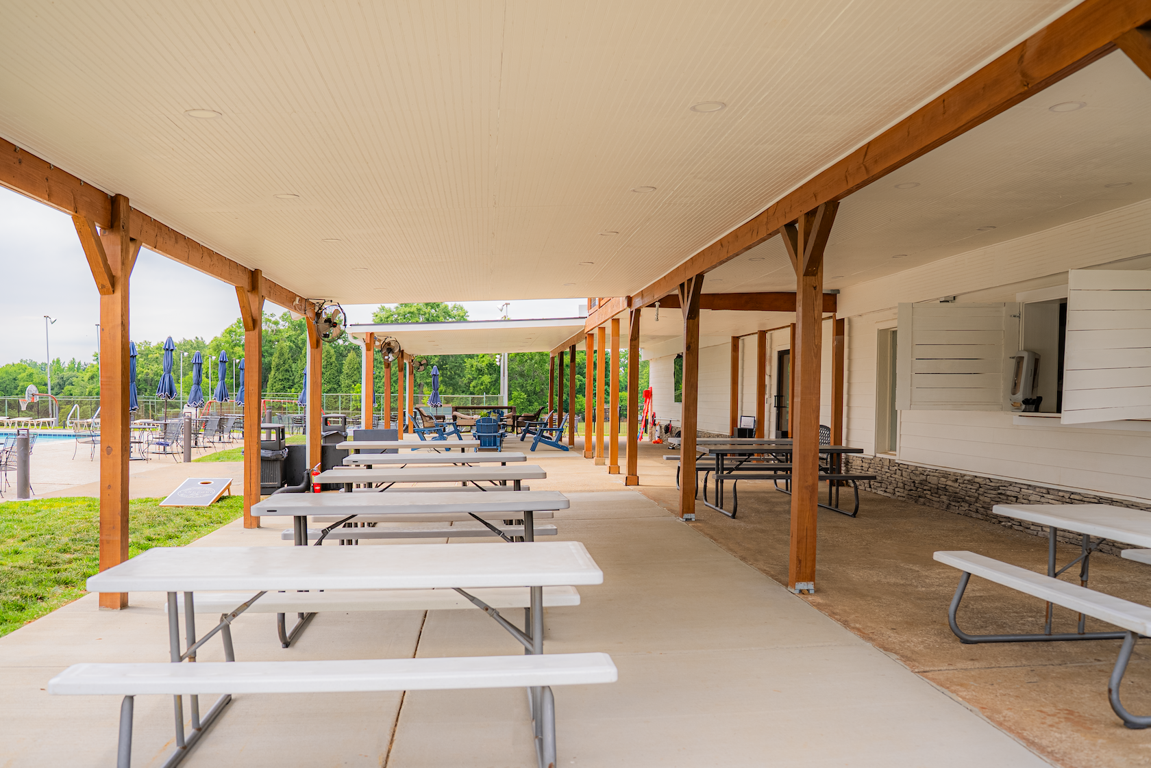 Empty outdoor patio area with picnic tables under a covered roof, overlooking a swimming pool with closed blue umbrellas, surrounded by a fence and green trees.