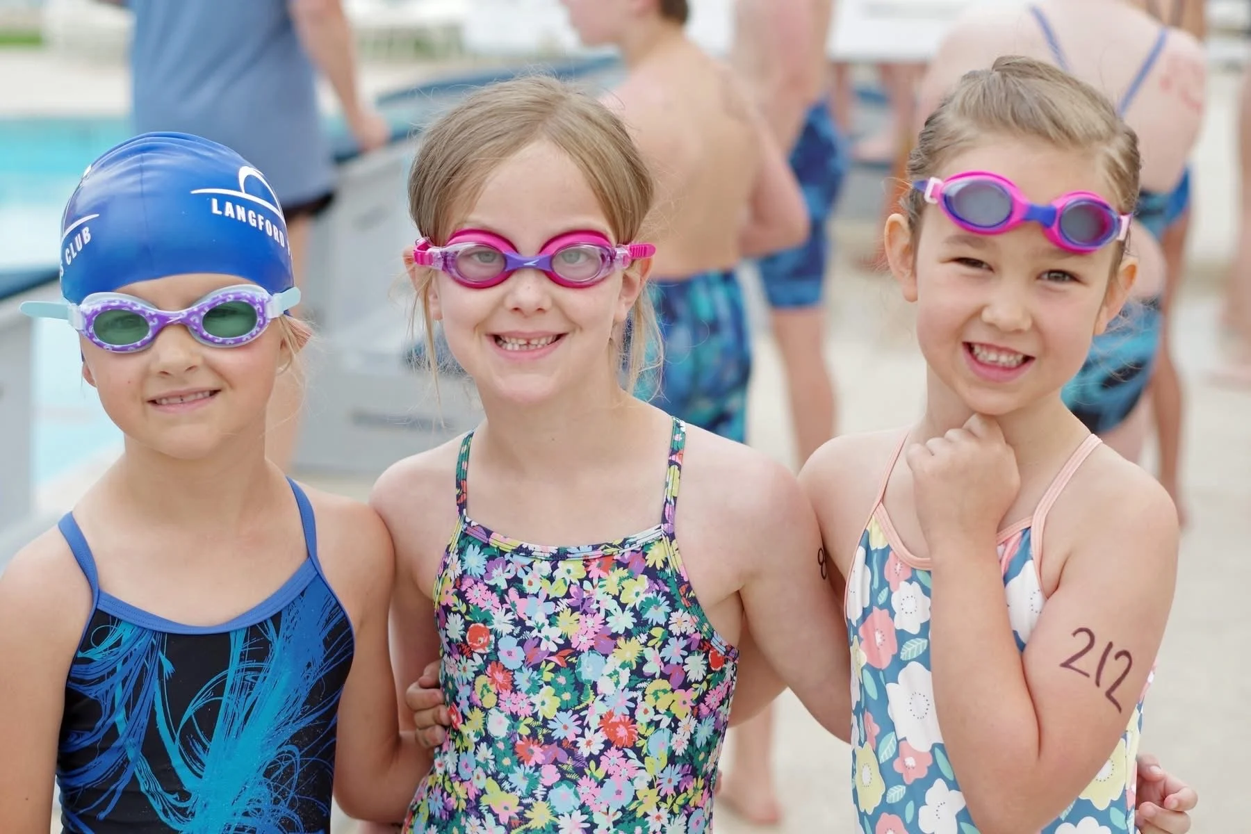 Three young girls in swimsuits and goggles smiling at a swimming pool, with other swimmers in the background.