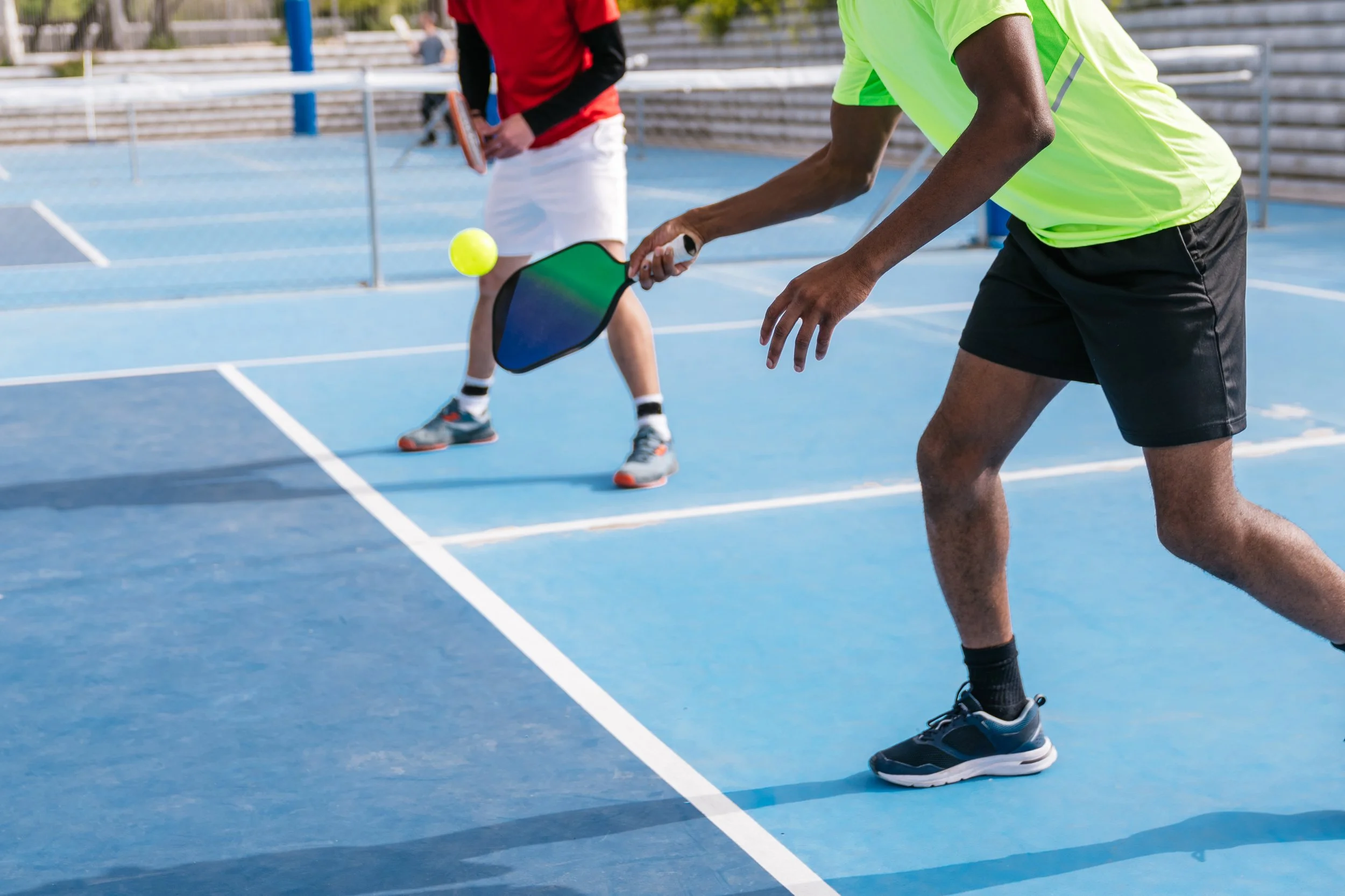 Two people playing pickleball on a blue outdoor court. One person is hitting a yellow ball with a paddle, while the other stands nearby.