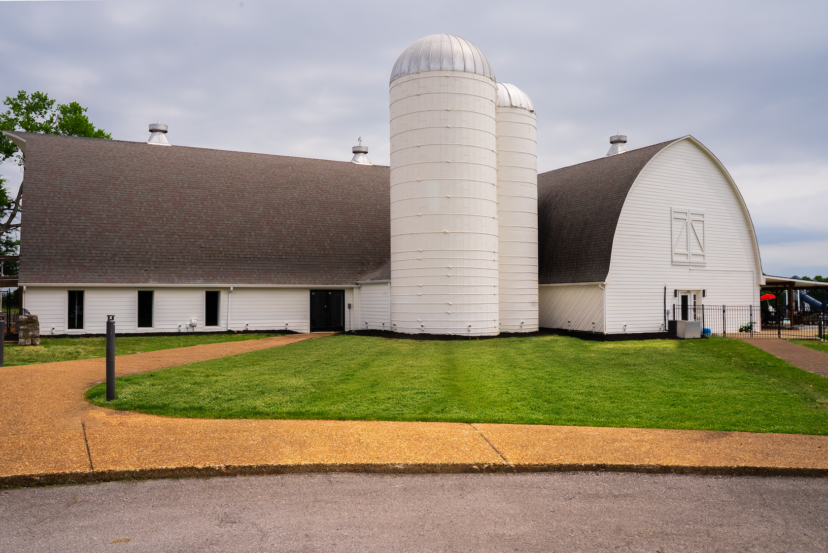A white barn with two silos, a grass lawn, and a paved walkway in front, under a cloudy sky.