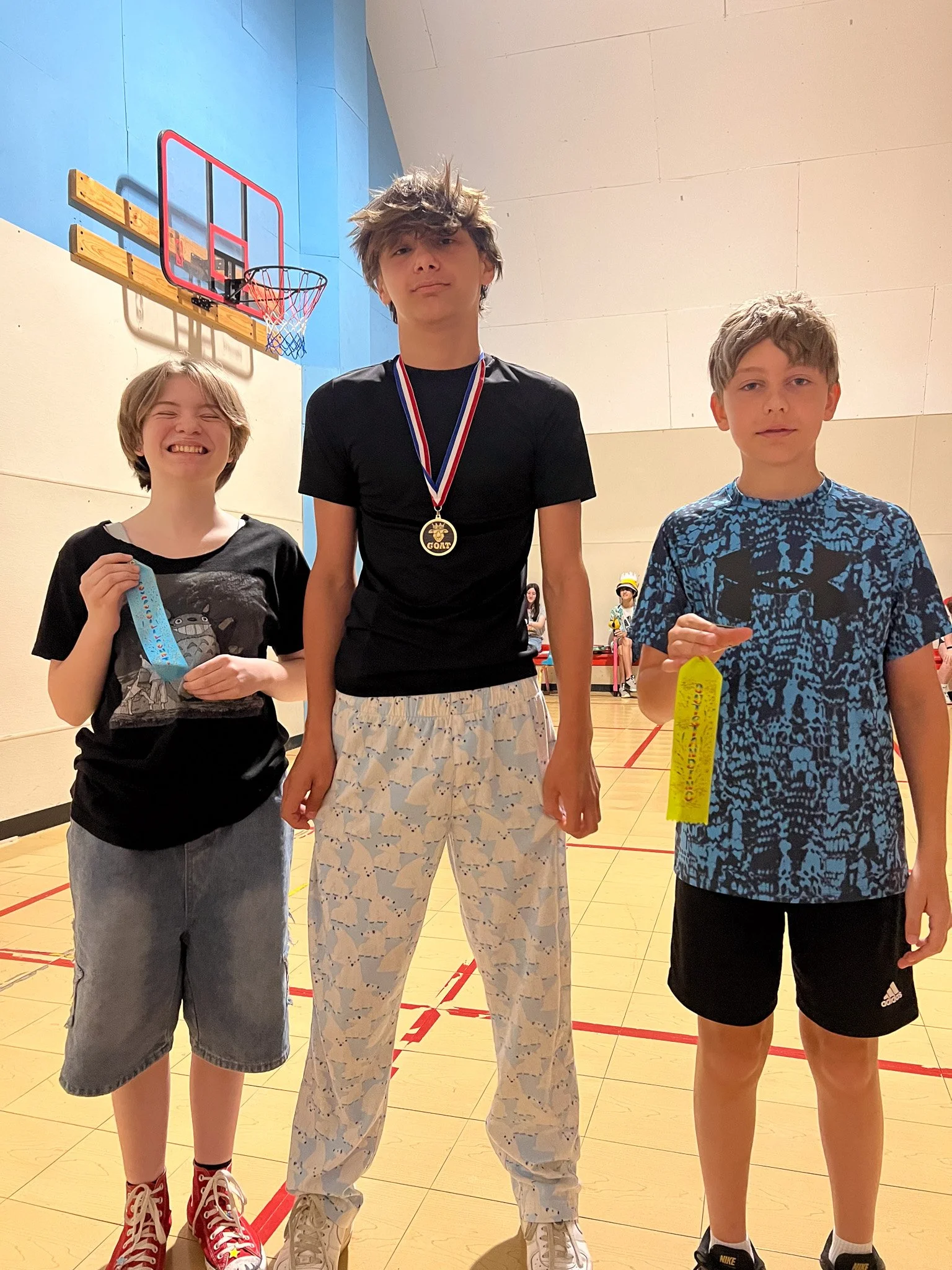 Three boys standing in a gymnasium, one wearing a medal around his neck and holding a ribbon, with a basketball hoop in the background.