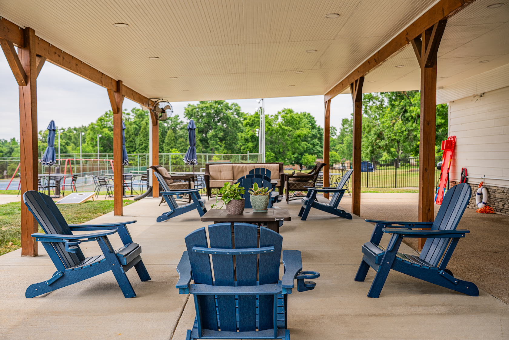 Covered outdoor patio with blue Adirondack chairs, a table with potted plants, a cushioned sofa, other chairs, and a view of a grassy area and tennis courts in the background.