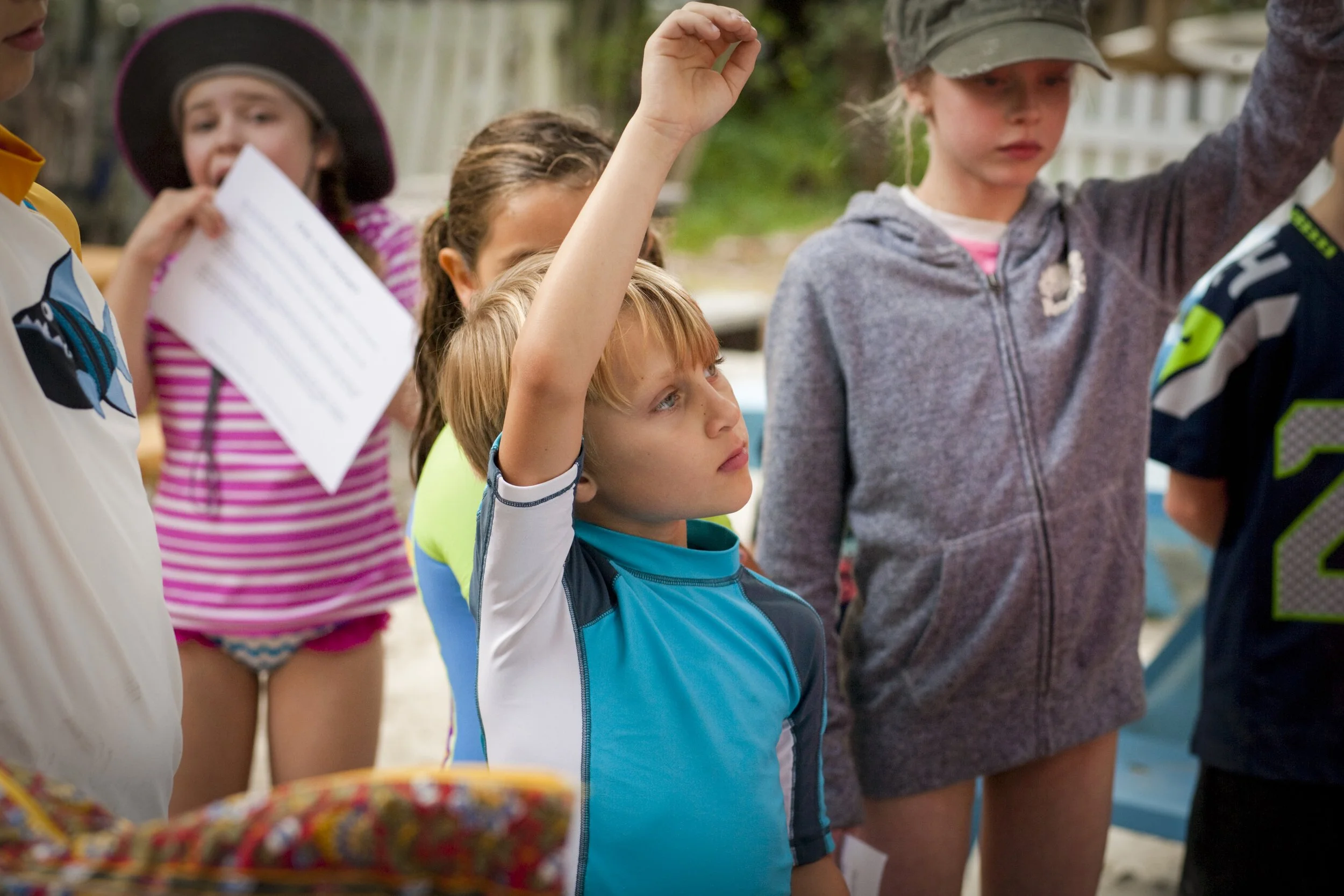 Group of children outdoors, with one boy raising his hand, some holding papers, dressed in casual and sports clothing.