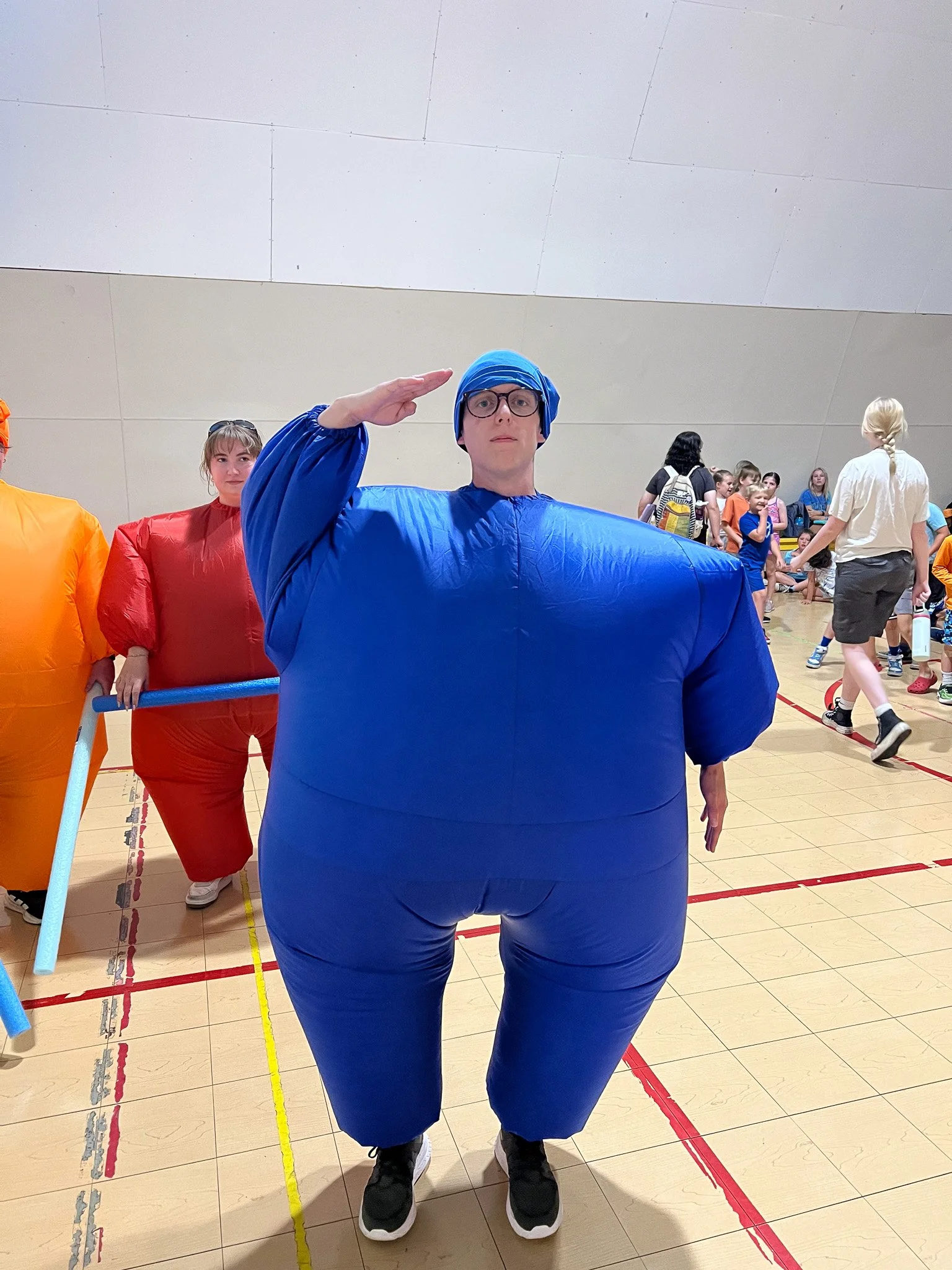 Person in a blue inflatable costume dressed as a character with glasses salutes at a costume event in a gymnasium with children and adults in the background.