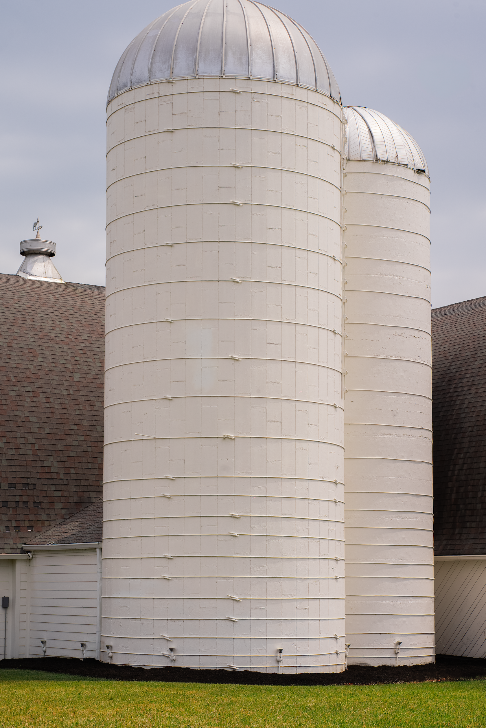 White barn with three silos and a gray shingled roof, green grass in front, and cloudy sky.