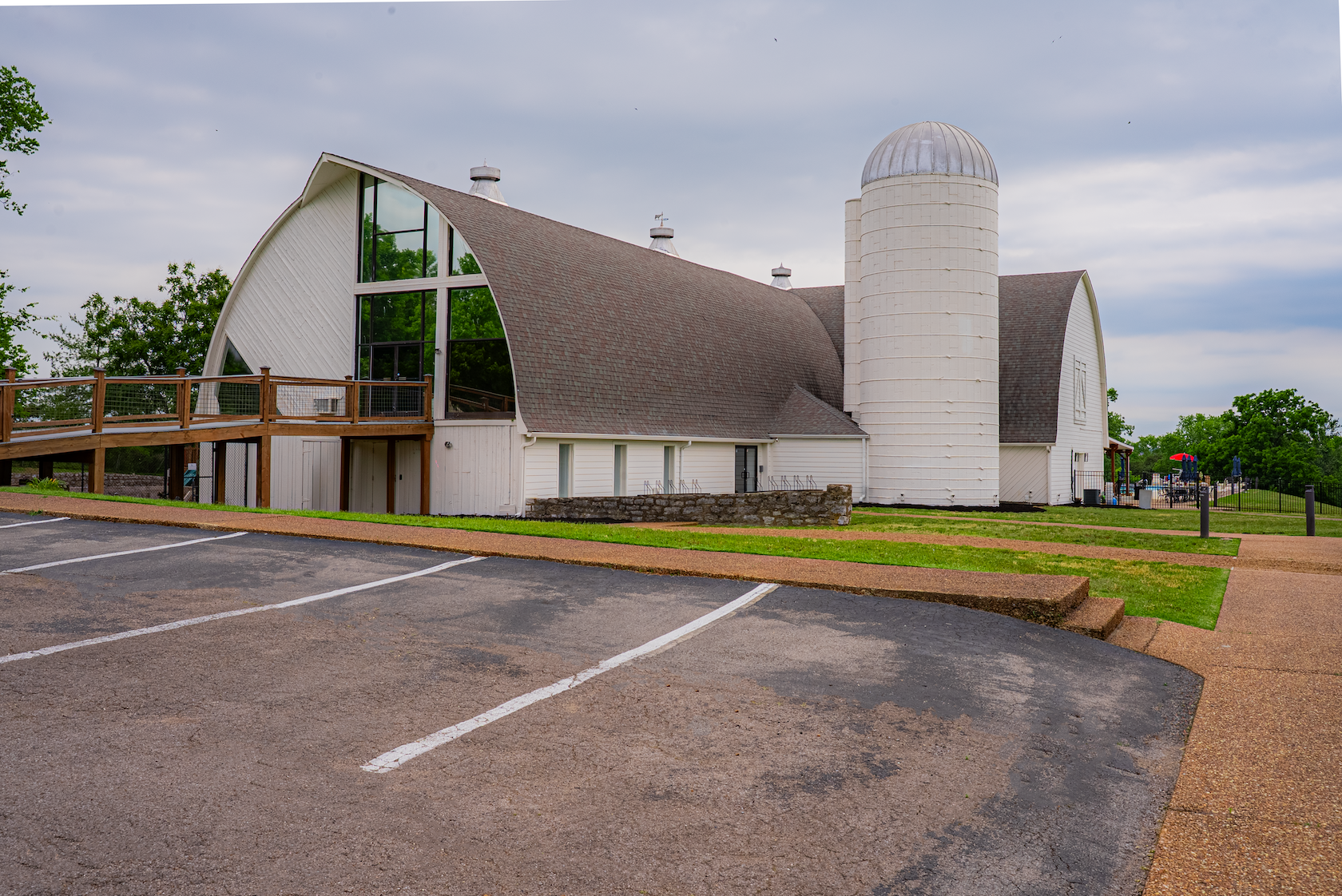 White building with a tall, rounded silo and arched roof, large windows, wooden walkway, parking lot in foreground, trees, and an outdoor area with umbrellas and seating in the background.