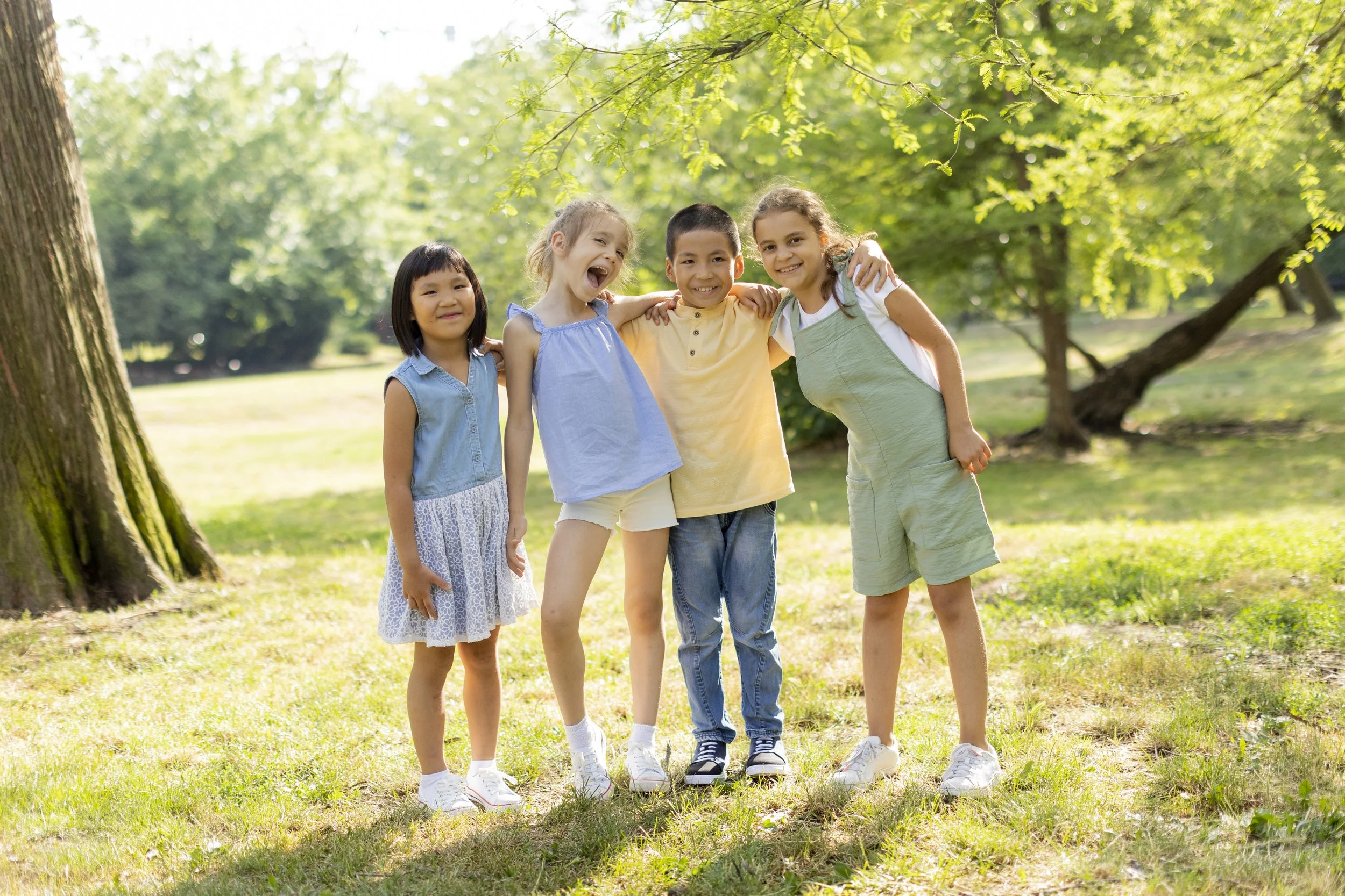 Group of four diverse children standing together outdoors in a park, smiling and hugging each other.