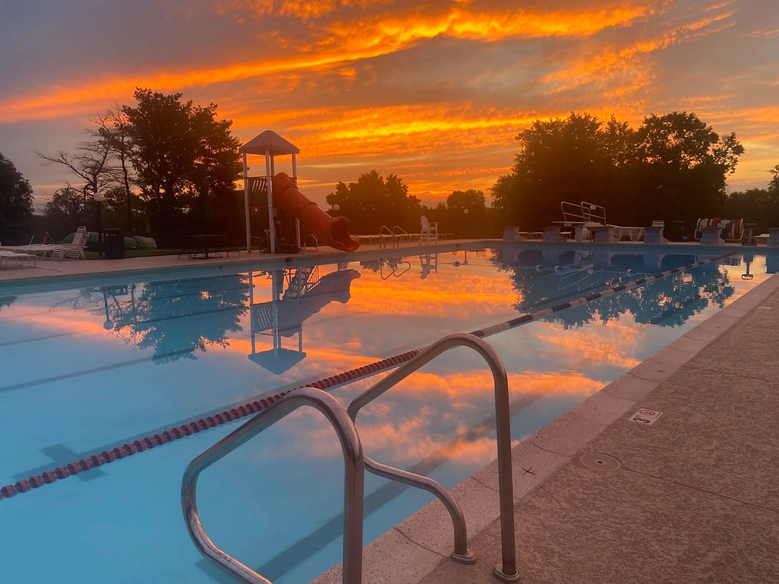 An outdoor swimming pool reflecting a vibrant sunset sky with orange and yellow clouds, surrounded by trees and poolside chairs.