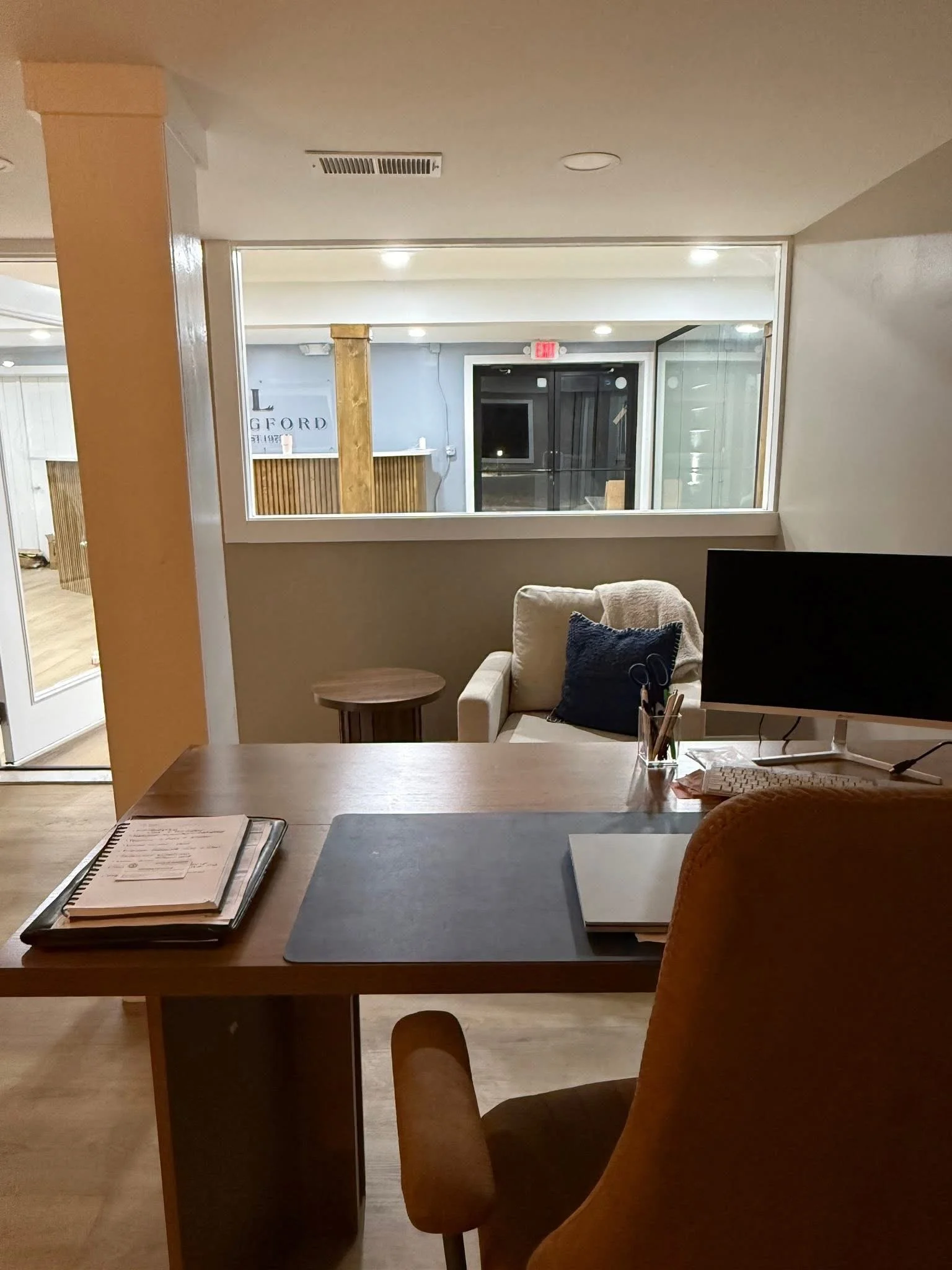 Office workspace with a wooden desk, a monitor, papers, and a chair in the foreground; behind is a sitting area with a beige armchair, a small side table, and a large window view of an outdoor area with a wooden fence and a sign partially visible, reading "L" and "FORD."