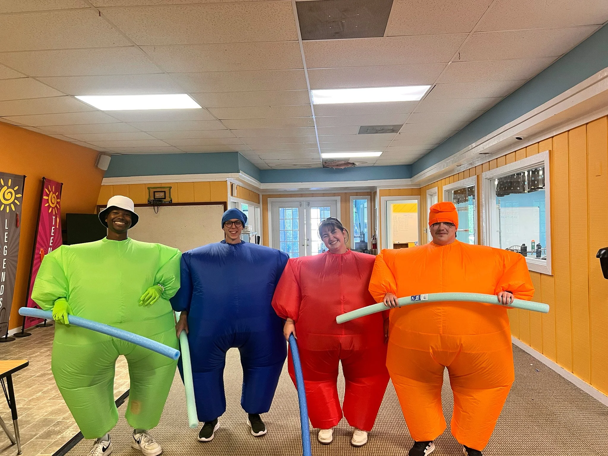 Four people dressed in oversized, colorful inflatable suits resembling popcorn containers, standing inside a room with bright yellow walls, windows, and banners.