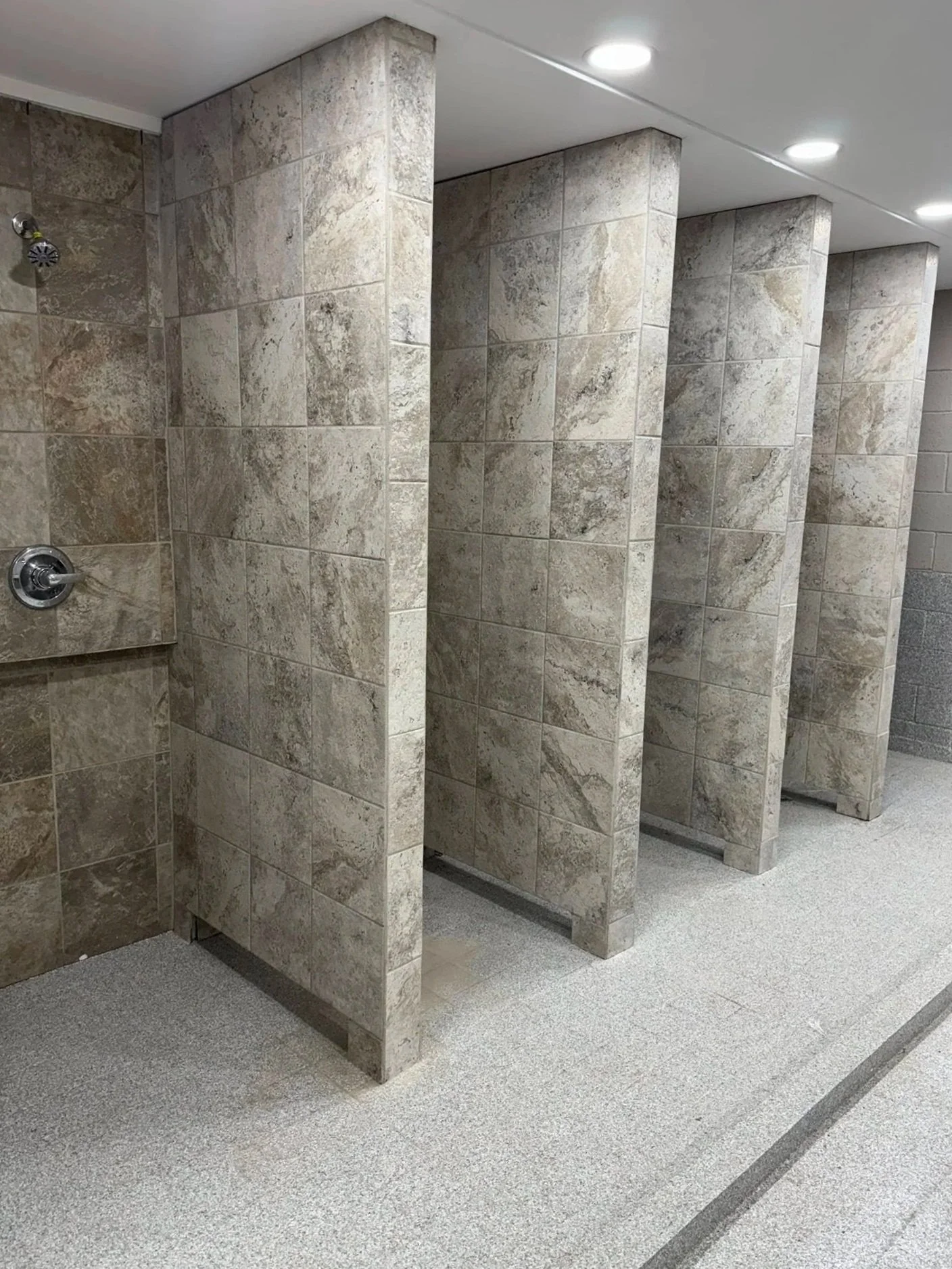 Public men's restroom with multiple tiled stalls and a urinal on the wall, illuminated by ceiling lights.