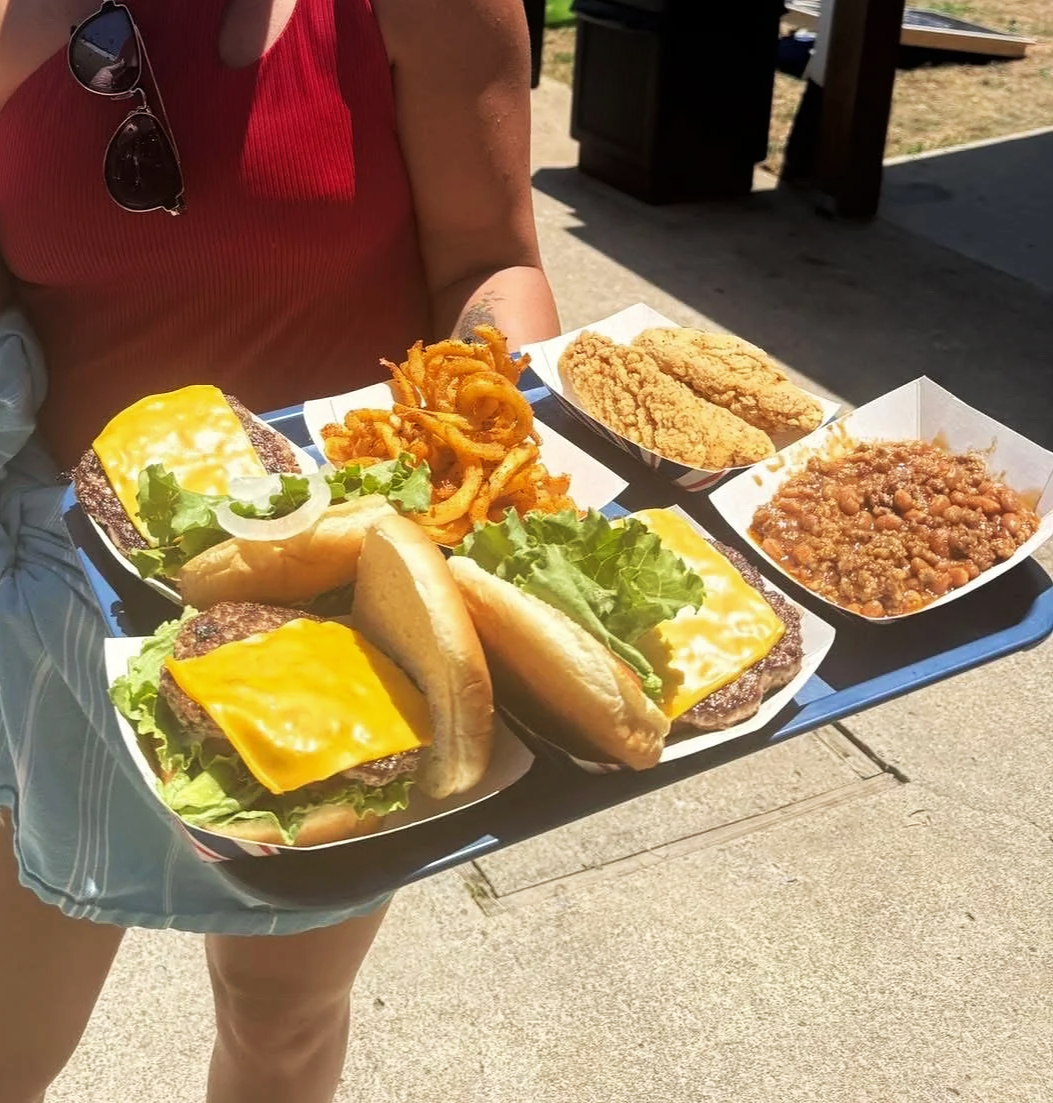 A person carrying a tray with cheeseburgers, fried calamari, fried chicken tenders, baked beans, and mashed potatoes with gravy.