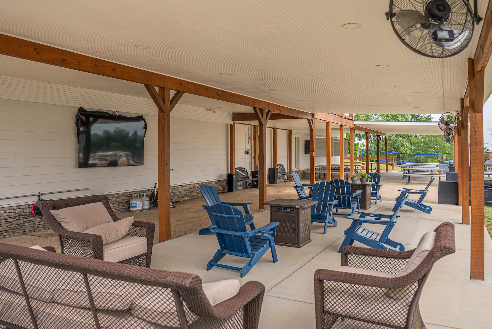 Outdoor covered patio with blue Adirondack chairs, wicker sofa with beige cushions, TV mounted on wall, ceiling fans, potted plants, and nearby picnic tables and playground in the background.