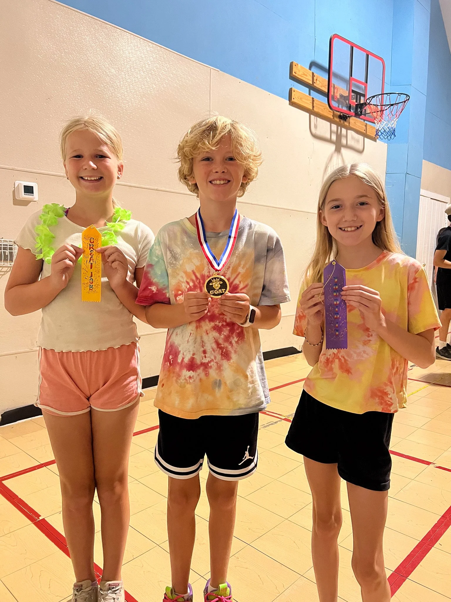 Three children standing on a gymnasium floor, holding colorful ribbons and a medal, smiling after a sports event.