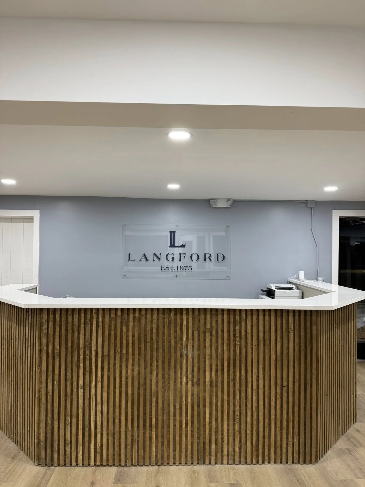 Reception desk with a white countertop and wooden slatted front in an office with a logo on the wall that reads "Langford EST. 1975".