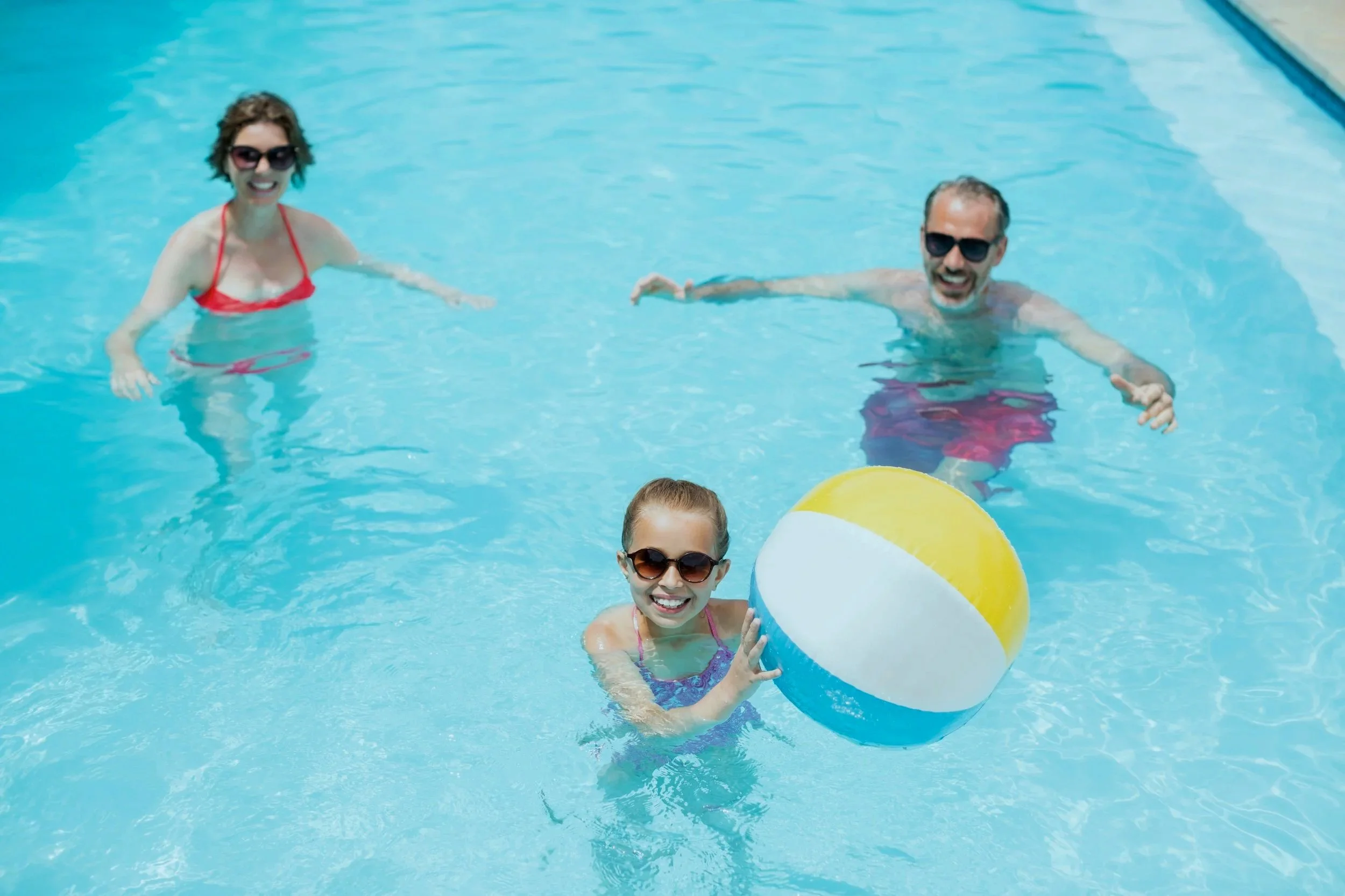 A family enjoying a swim in a pool with a colorful beach ball, all wearing sunglasses and smiling.