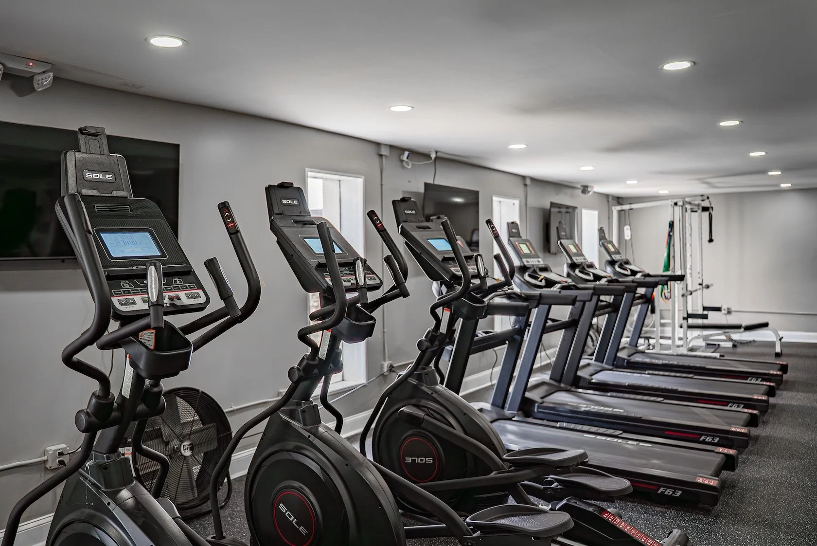 Row of exercise machines including elliptical trainers, upright bikes, and treadmills in a well-lit gym with gray walls and windows.