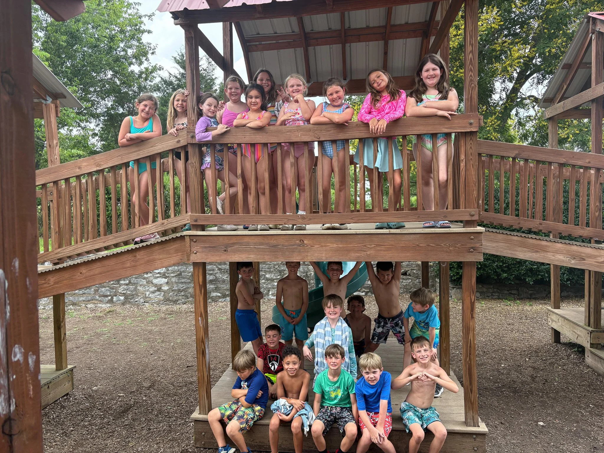 Group of children on a two-level wooden playground structure, smiling and posing for the camera. The kids are dressed in summer clothes and swimwear, standing on the upper deck and sitting or standing underneath.