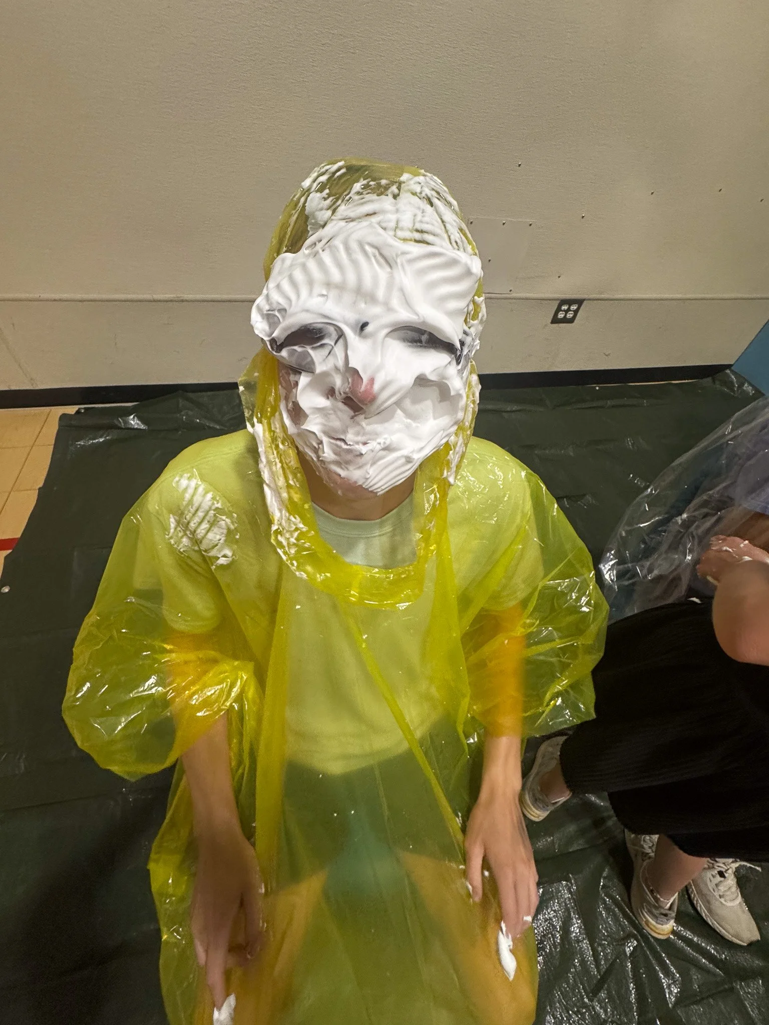 Person sitting on the floor wearing a yellow plastic poncho and a mask made of whipped cream and other materials, resembling a face with exaggerated features.