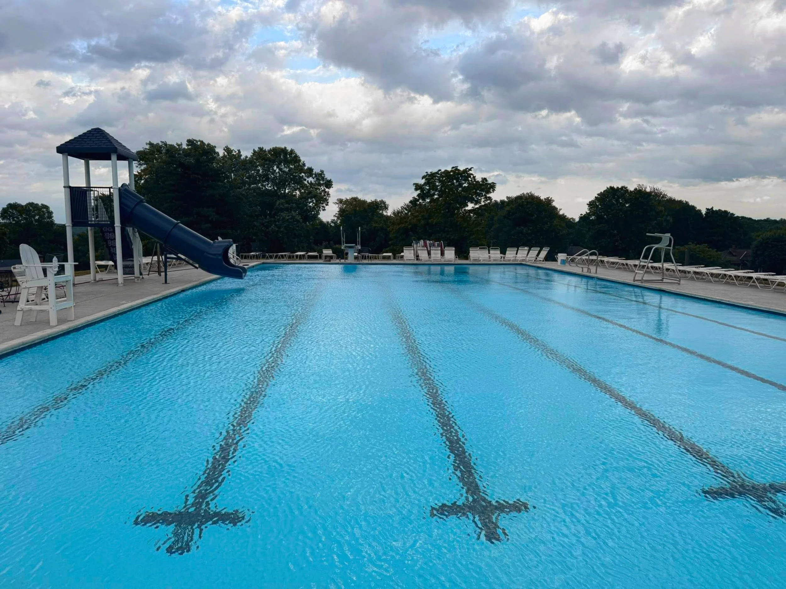 Empty outdoor swimming pool with lane lines, slides, and lounge chairs on a deck, surrounded by trees and cloudy sky.