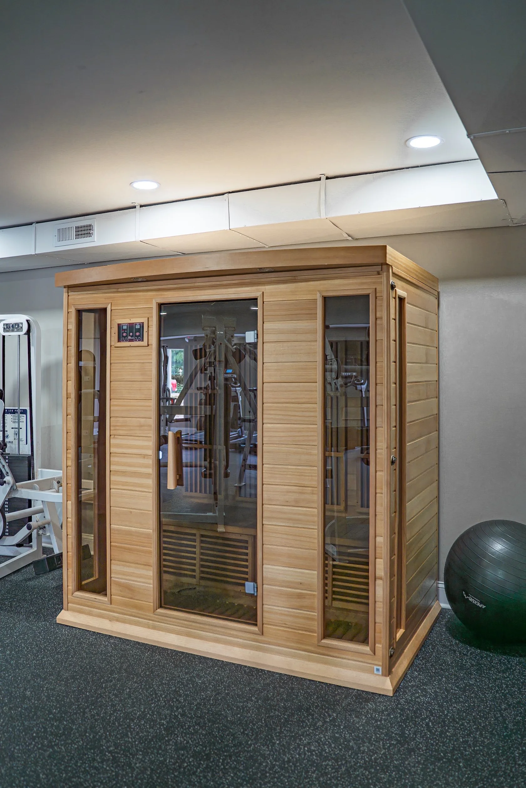 A wooden infrared sauna in a gym or wellness center setting, with glass doors and side windows, surrounded by gym equipment and a large exercise ball on a dark speckled floor.