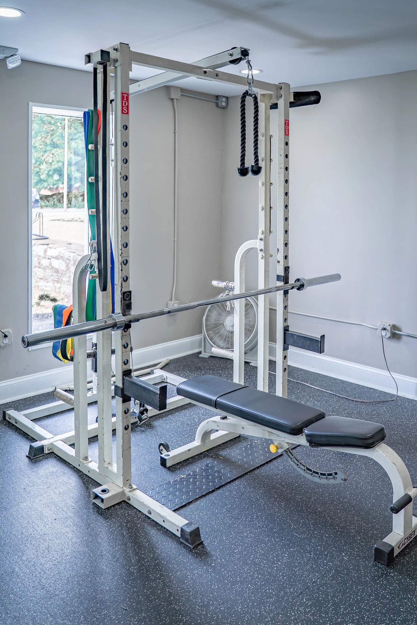 Home gym with weightlifting bench, resistance bands hanging on a frame, fan near the wall, and window with trees outside.