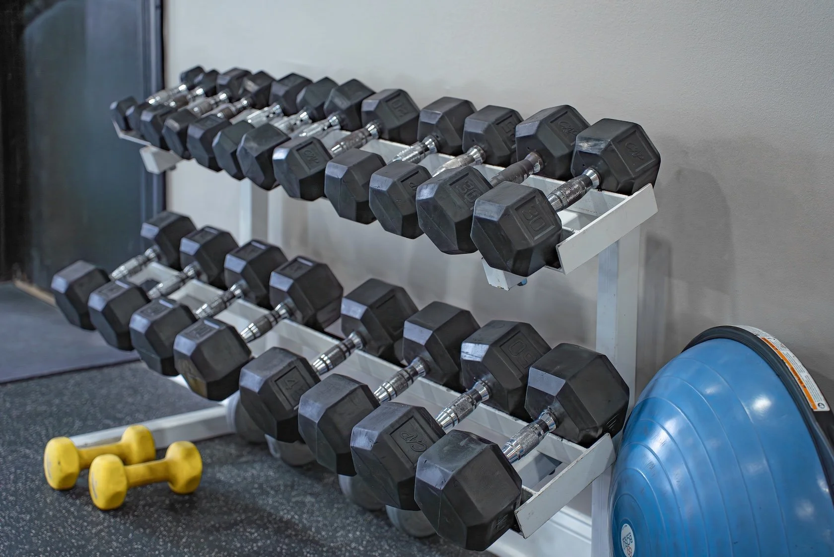 A set of black dumbbells on a white rack in a gym, with yellow bell-shaped weights on the floor and blue exercise balls to the side.