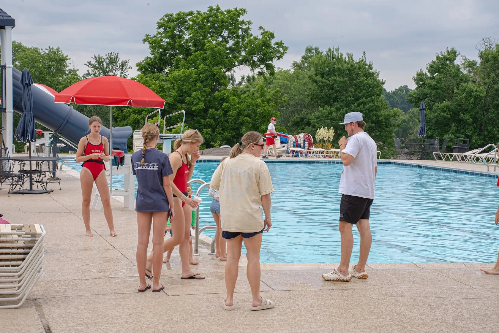 People gather at the edge of a swimming pool, preparing to swim. There is a lifeguard on duty, and a slide in the background. The scene is outdoors on a cloudy day, with lush green trees surrounding the pool area.