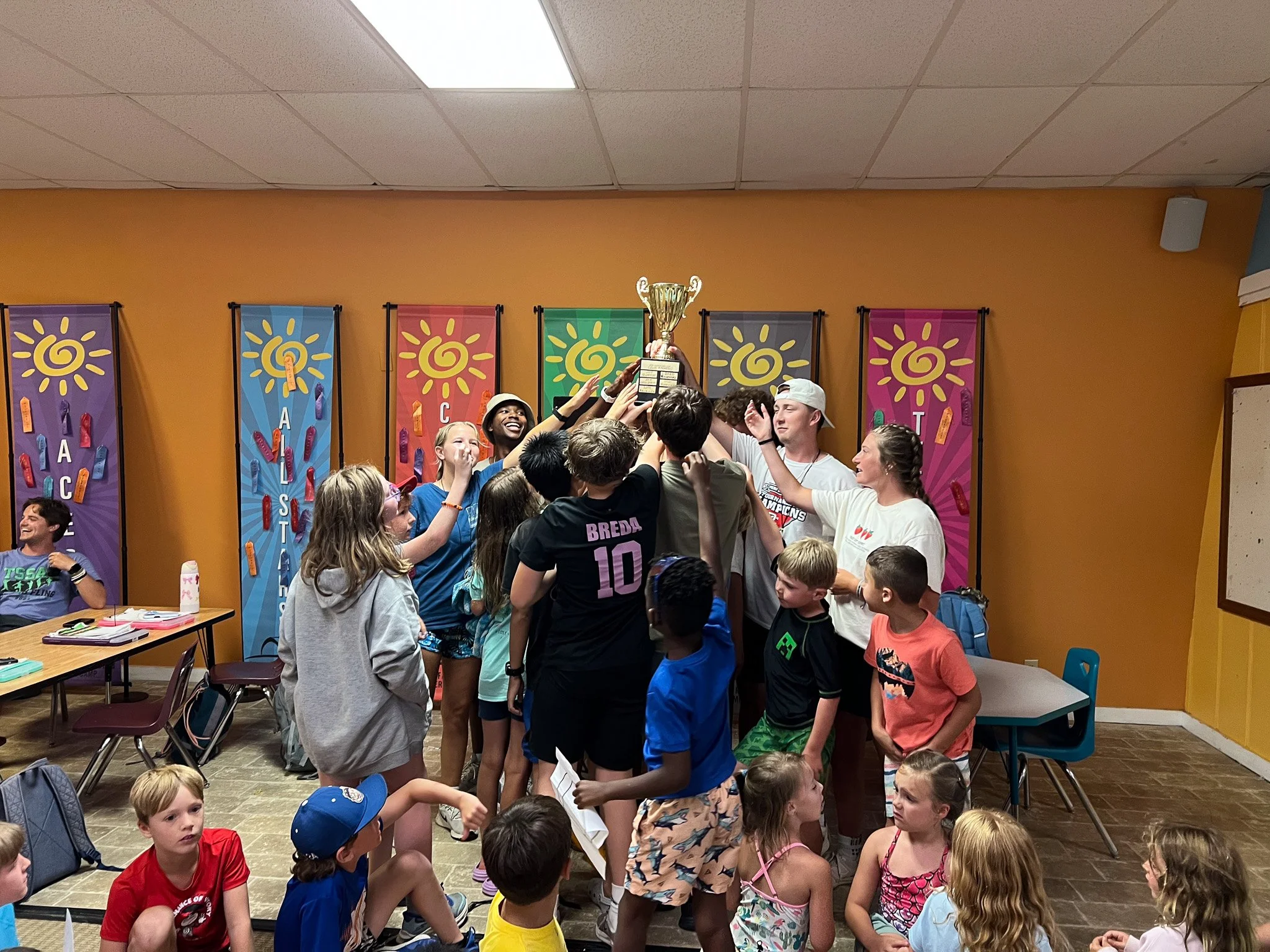 A group of children and teenagers celebrating victory as they lift a trophy together in a colorful indoor setting, with some kids sitting on the floor and others standing around.