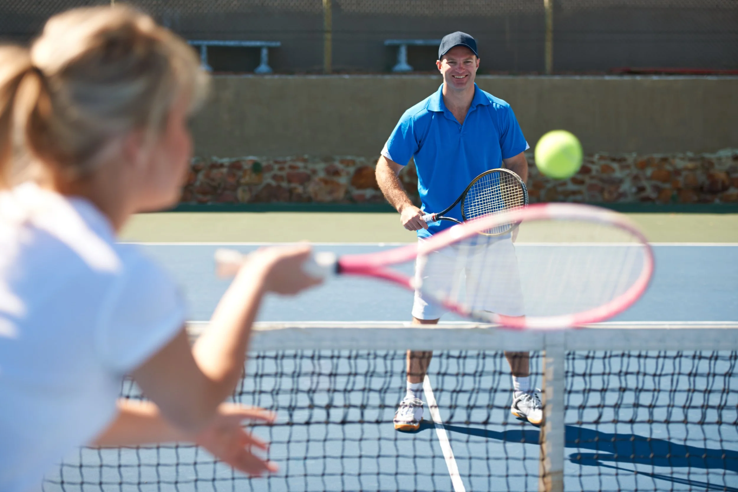 A young girl playing tennis on an outdoor court with a man in the background preparing to hit a tennis ball.