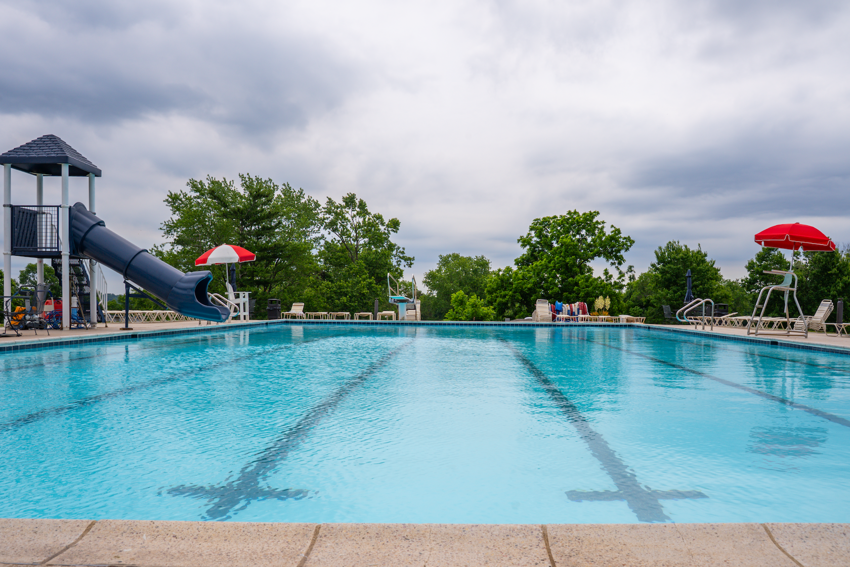Empty outdoor swimming pool with water slide, surrounded by lounge chairs and red umbrellas, with green trees and cloudy sky in the background.