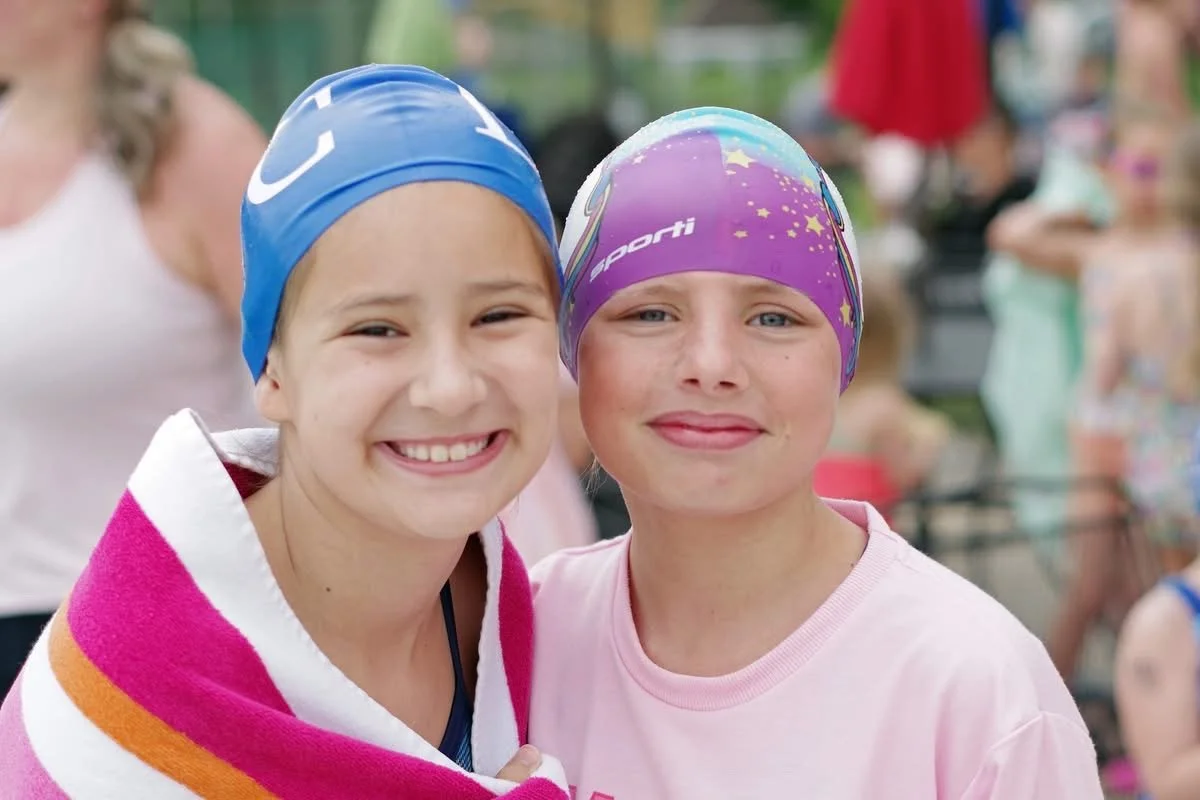 Two young girls wearing swim caps and pink t-shirts smiling at the camera at a swimming event.