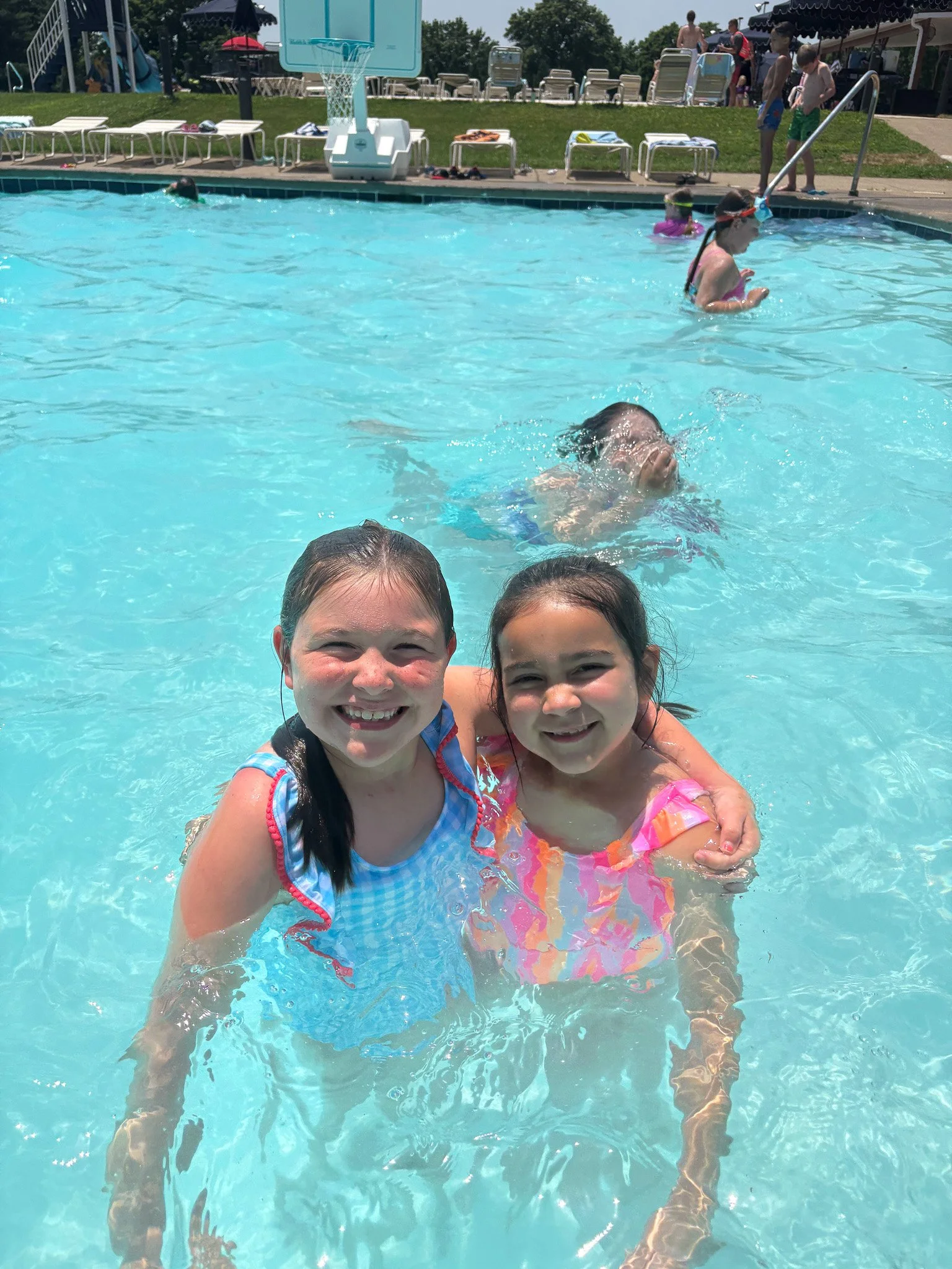 Two young girls smiling and hugging in a swimming pool on a sunny day, with other children swimming and playing in the background.