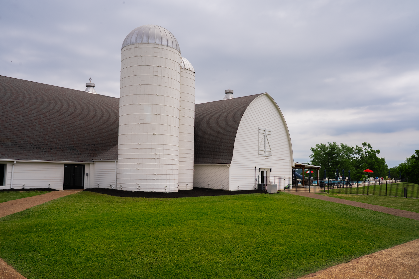 White barn with two silos, a paved walkway, green lawn, fence, and a swimming pool area with umbrellas and chairs in the background under a cloudy sky.