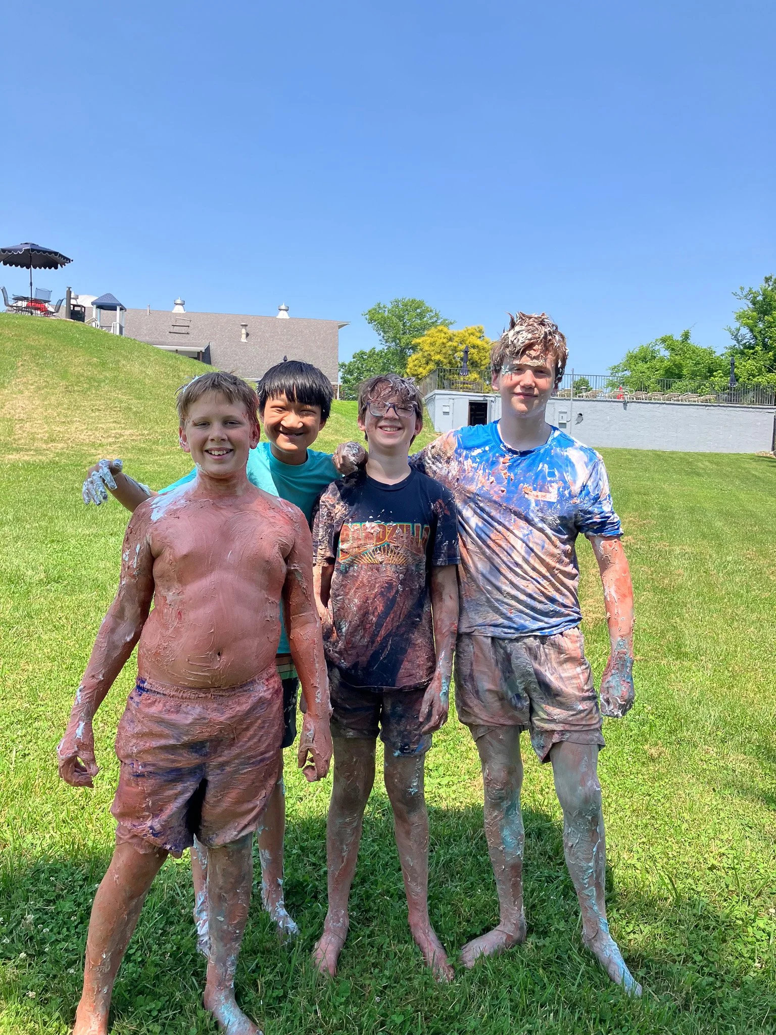 Four children covered in mud standing on grass with a house and trees in the background on a sunny day.