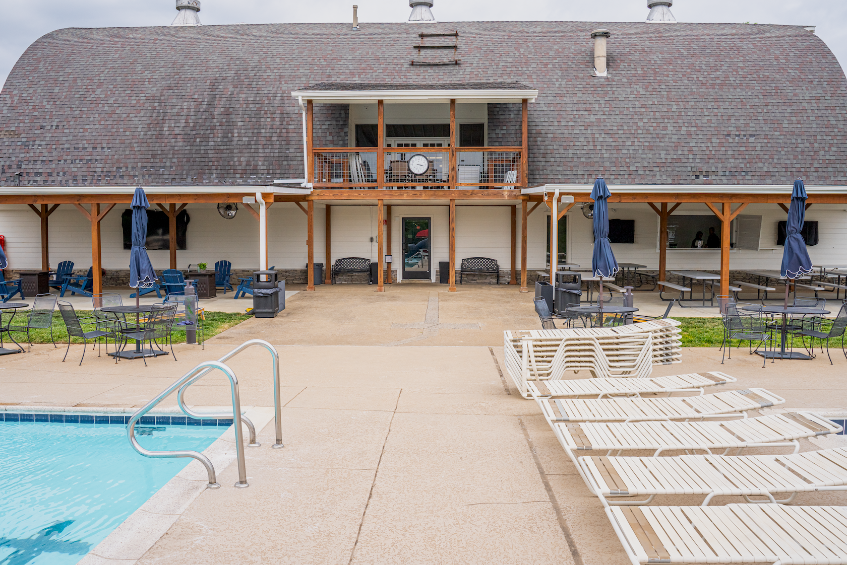 Outdoor swimming pool area with lounge chairs, tables, and umbrellas. A building with a wooden balcony and an outdoor dining area.