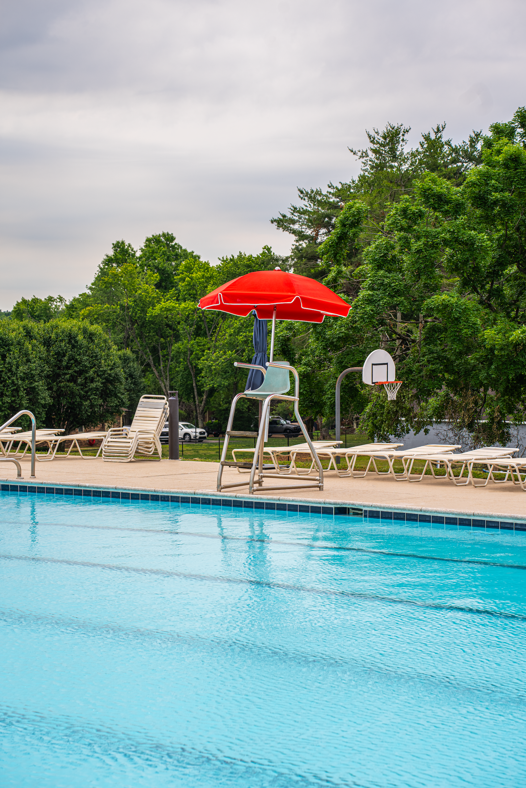 Empty outdoor swimming pool with lounge chairs, a red umbrella, a volleyball hoop, and trees in the background.