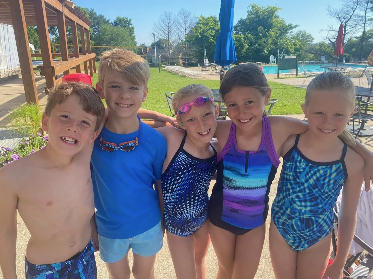 Group of five children in swimsuits smiling and posing together near a swimming pool on a sunny day.