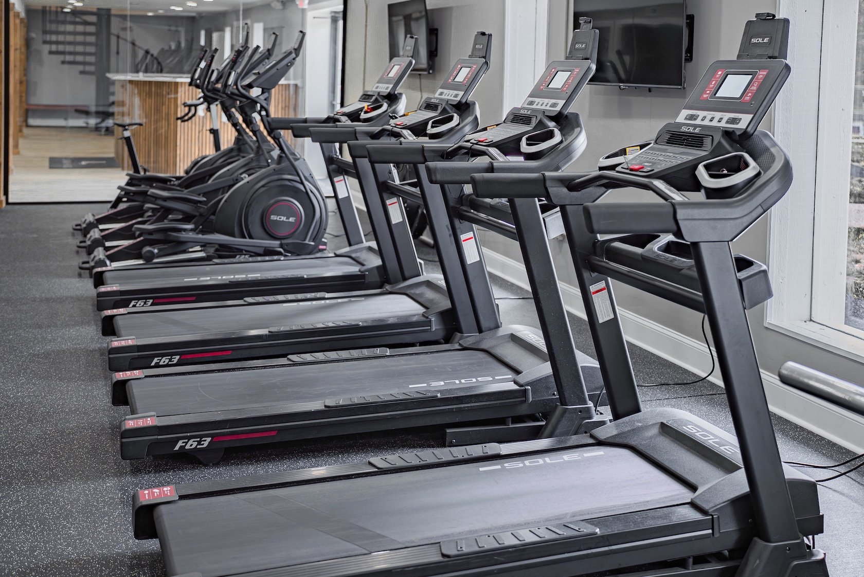 Row of six treadmills in a gym, with elliptical machines behind them and a television mounted on the wall.
