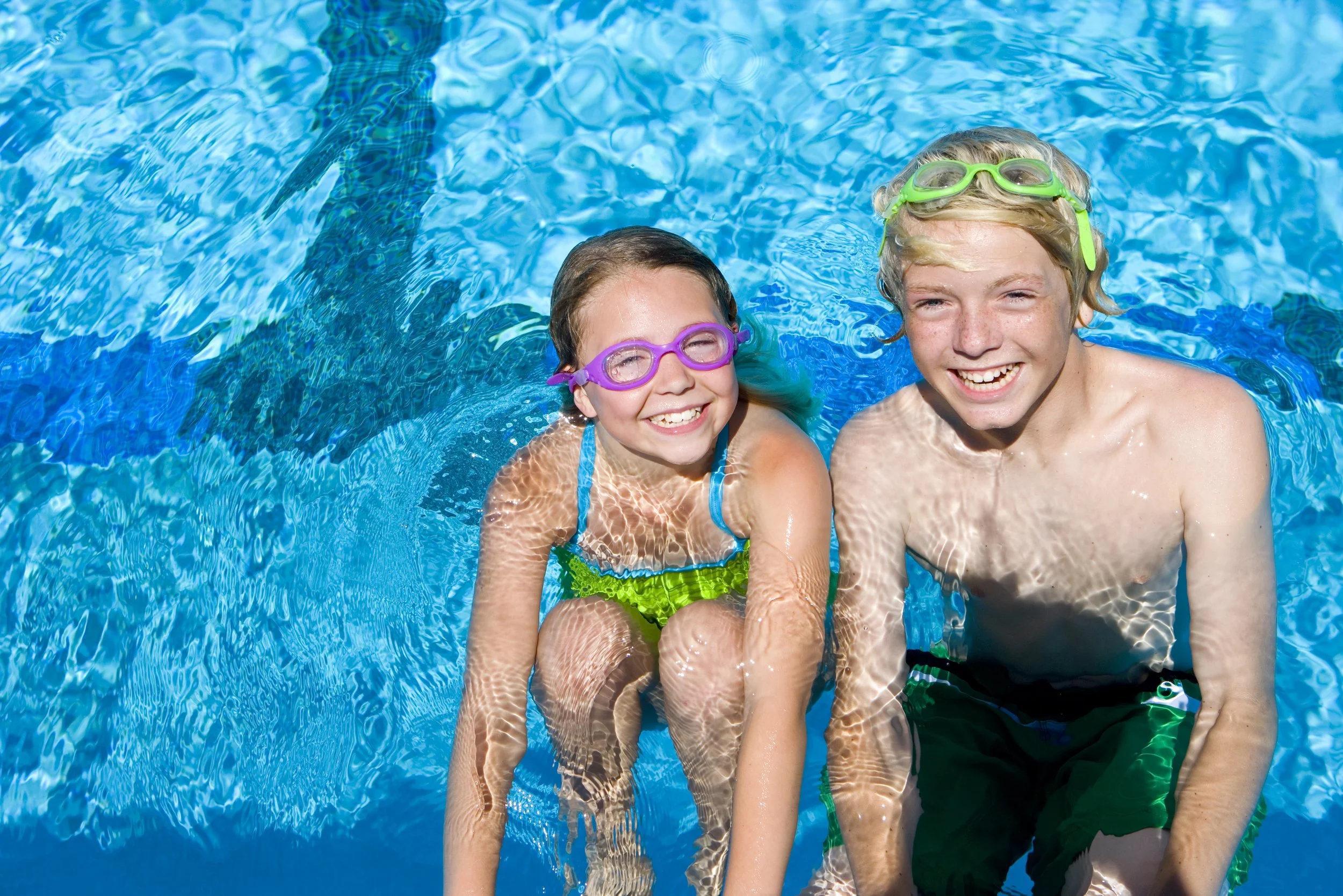 Two children, a girl and a boy, are smiling and sitting in a swimming pool. They are wearing colorful goggles and swimwear, enjoying a sunny day in the water.