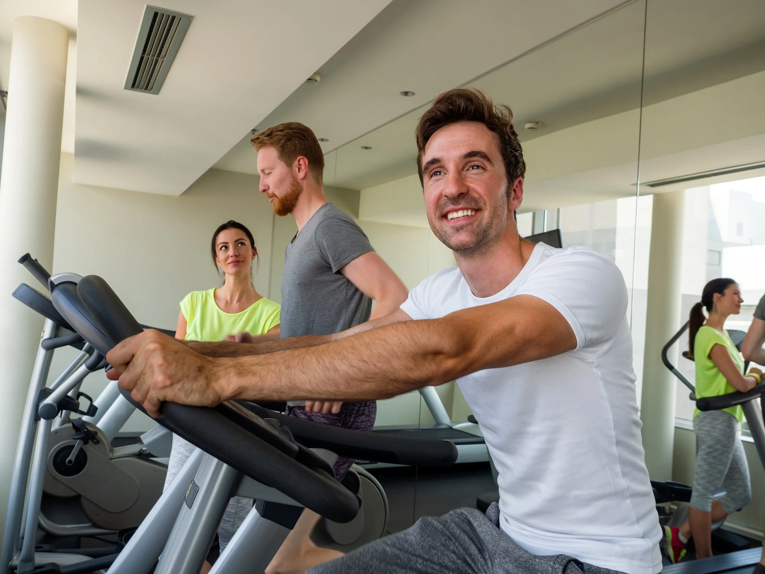 Man smiling on treadmill at gym, with two women and a man exercising in the background.