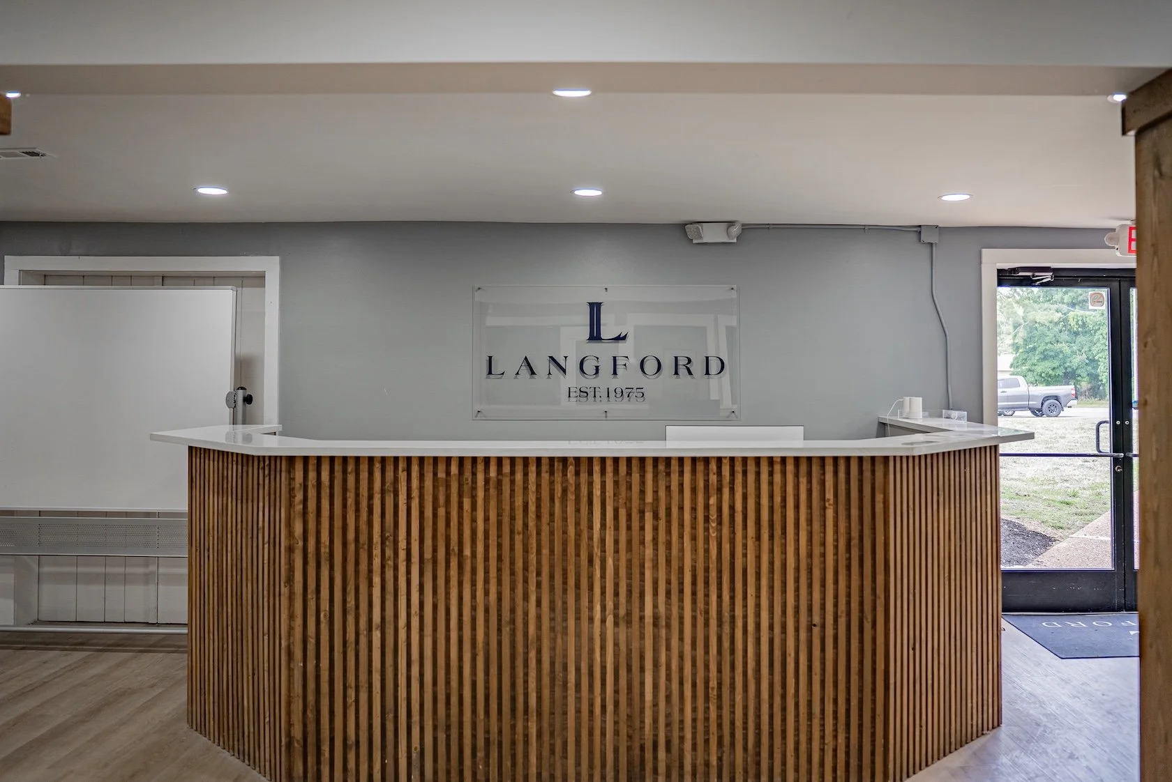 Reception area with a curved wooden counter and a sign that reads 'Langford Established 1975' on a wall behind it. There is a door to the outside with a vehicle visible through the glass.