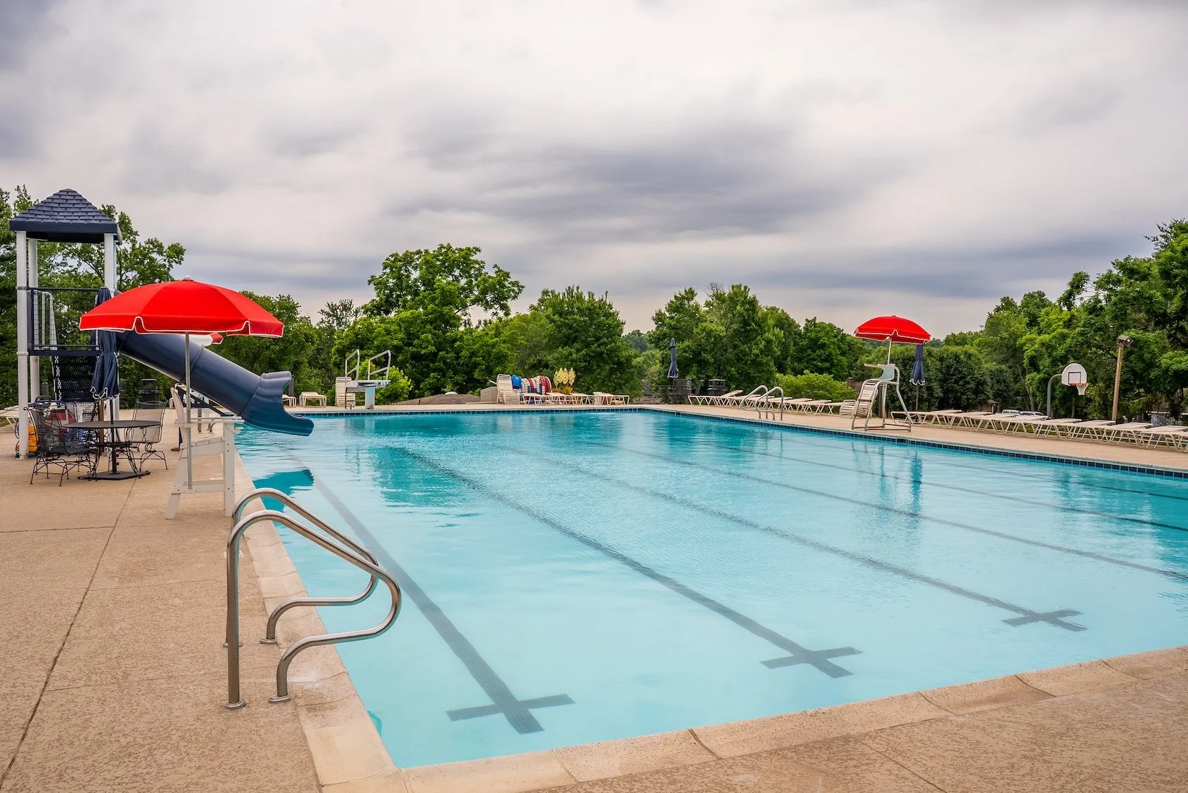 Empty outdoor swimming pool with a water slide, red umbrellas, and lounge chairs, surrounded by green trees and an overcast sky.