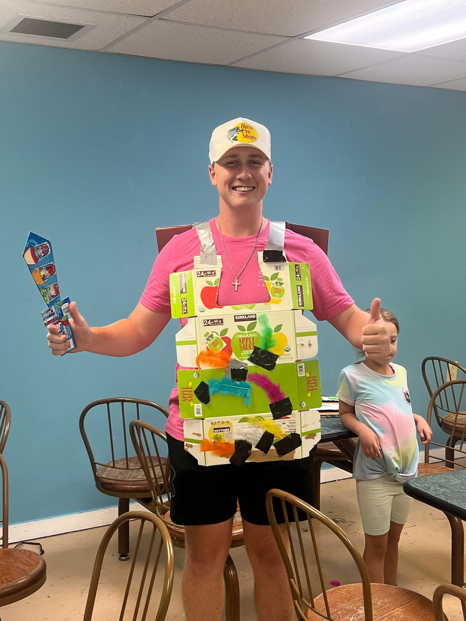 A young man smiling and giving a thumbs up is dressed in a costume made of apple juice boxes, colorful feathers, and duct tape, standing next to a young girl in a tie-dye shirt. They are in a room with a blue wall and wooden chairs.